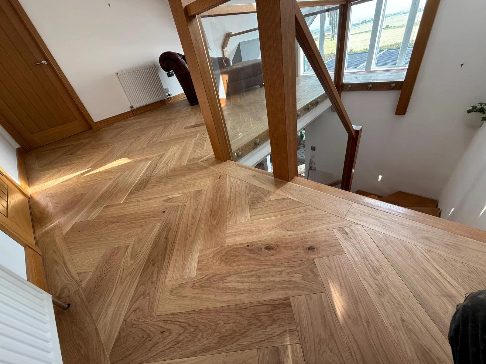 Wooden flooring with a chevron and herringbone pattern on the staircase landing and steps, with large windows visible in the background.