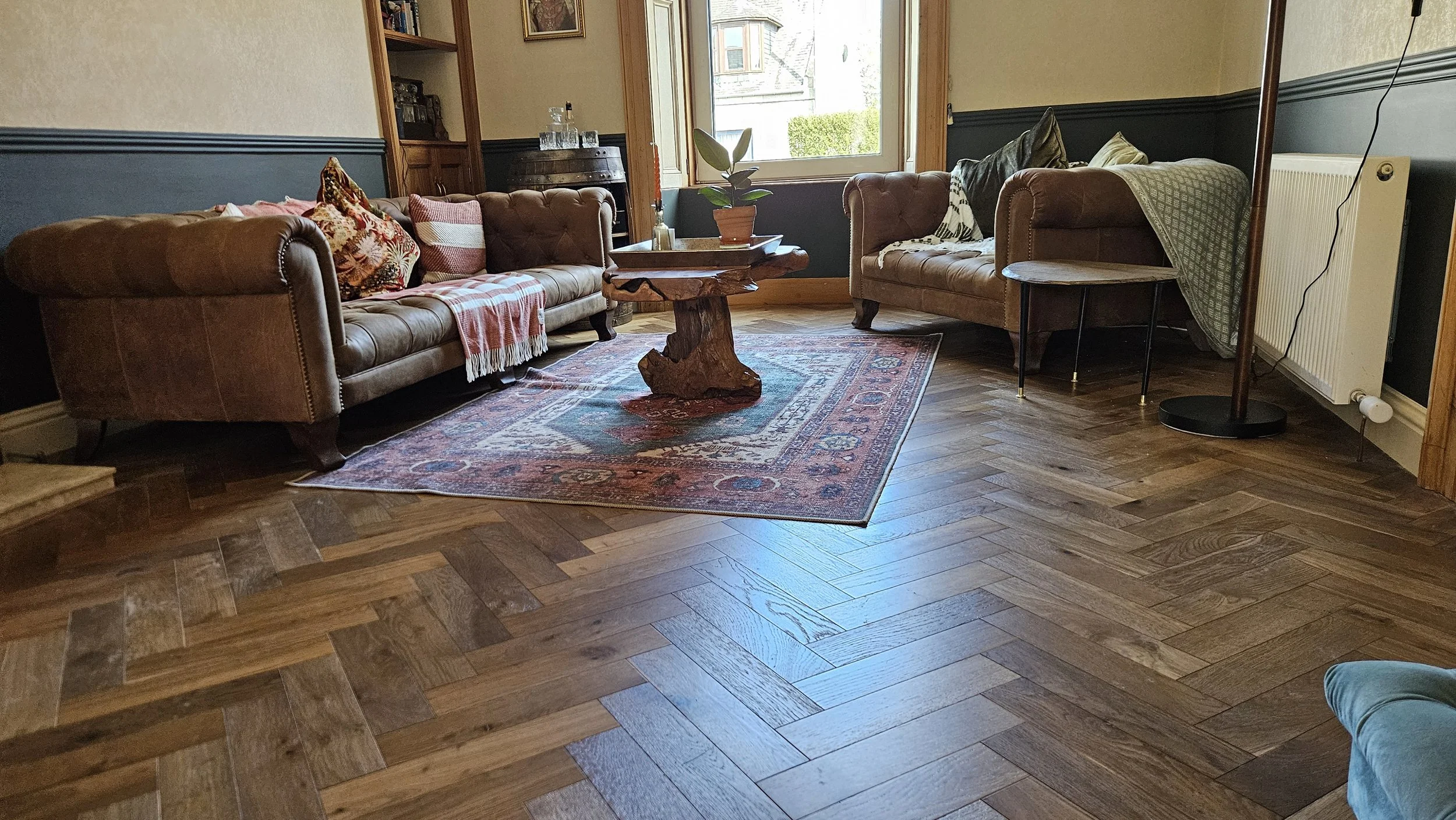 A living room with two brown leather sofas, a wooden coffee table with a potted plant, decorative pillows, a patterned rug, hardwood floor, large window, and a standing lamp.