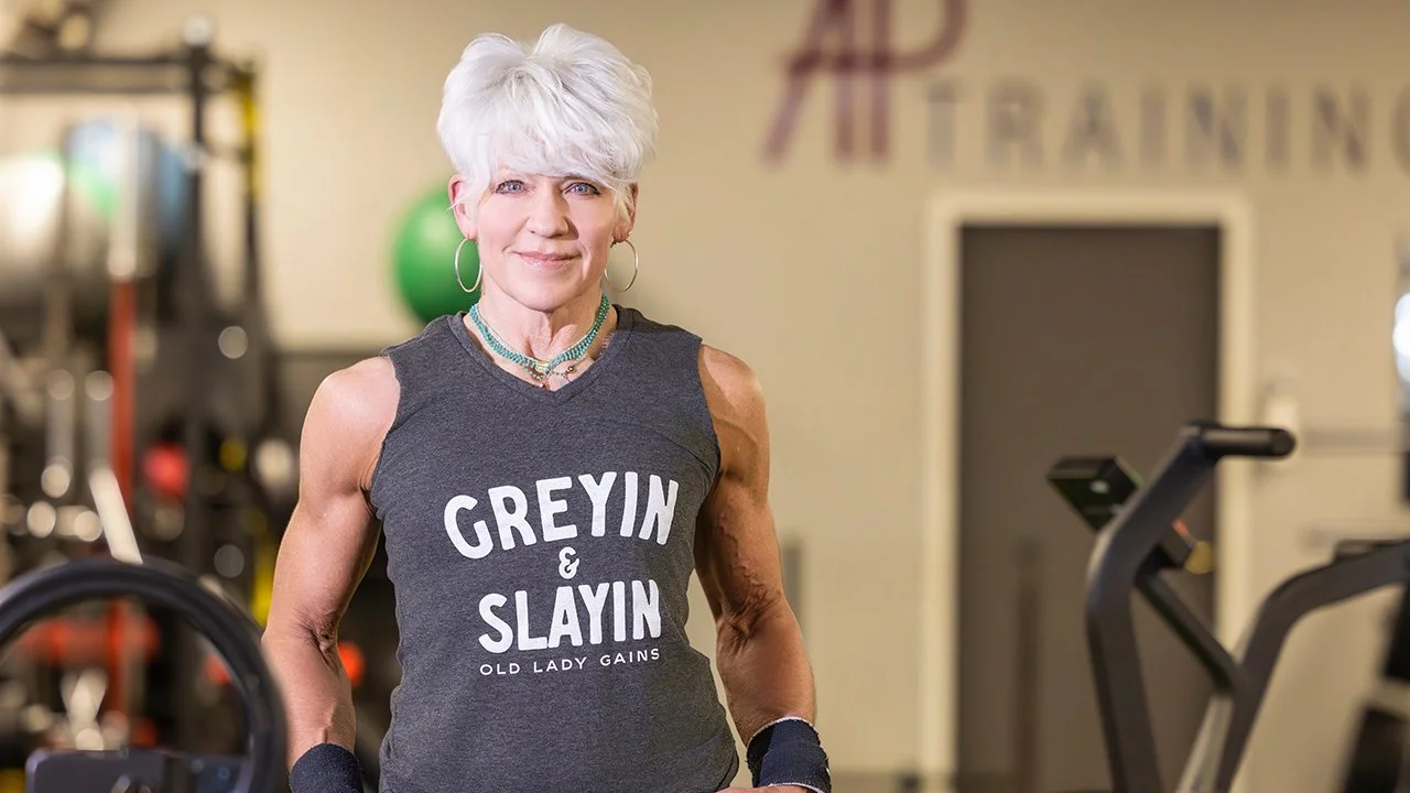 An older woman with short, white hair at the gym, wearing a sleeveless dark gray top that says 'GREYIN & SLAYIN OLD LADY GAINS'. She has hoop earrings, necklaces, and workout gloves, smiling at the camera with gym equipment in the background.