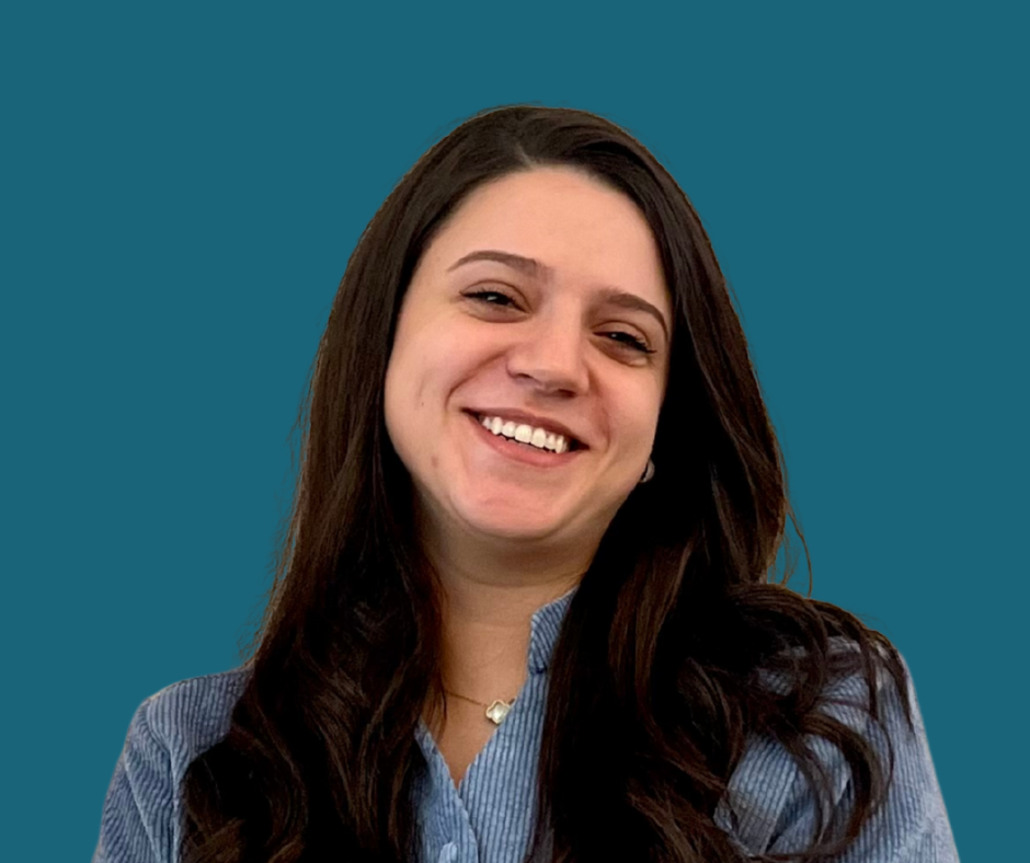 A woman with long dark hair smiling at the camera, wearing a gray blazer over a blue shirt, with a solid teal background.