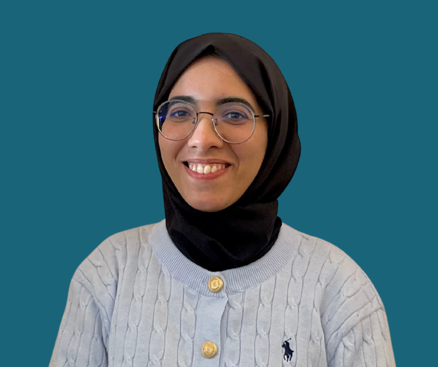 A woman smiling, wearing glasses, a black hijab, and a light-colored sweater with gold buttons and a small logo on the chest, against a teal background.