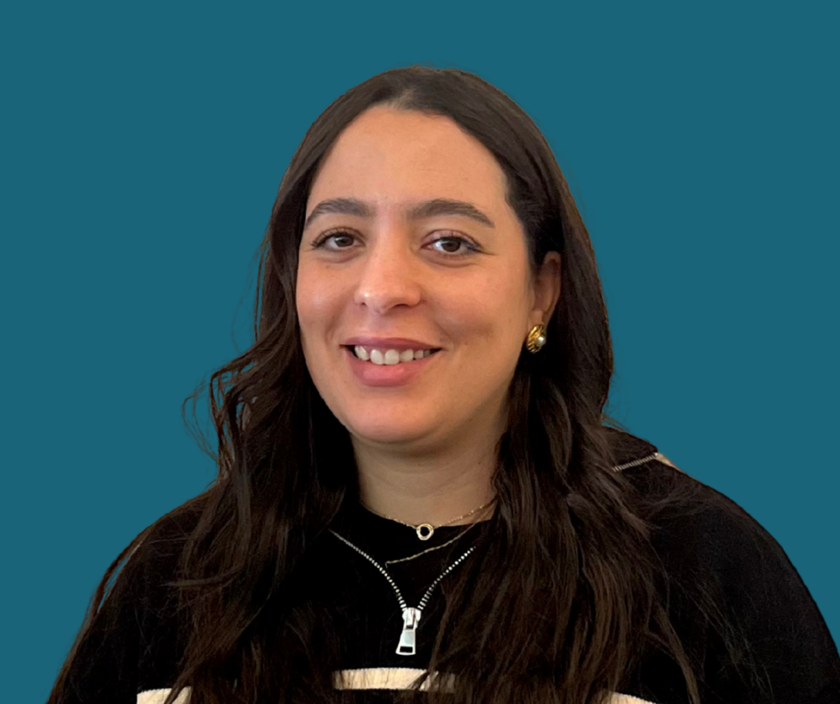 A young woman with long dark hair smiling, wearing a black top with a zipper and gold earrings, against a blue background.