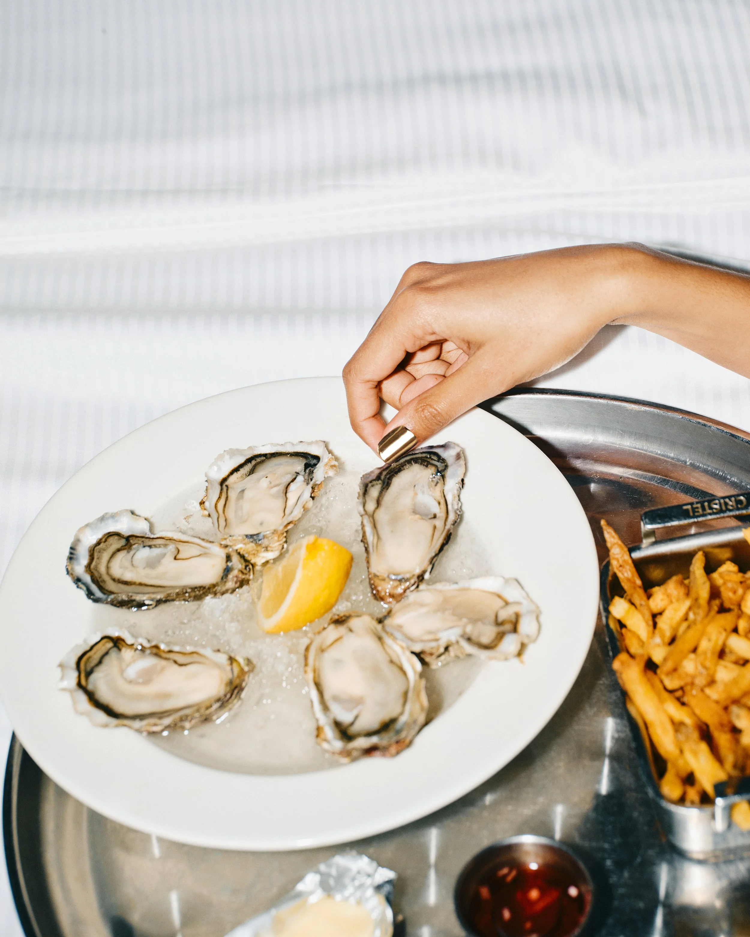 Hand wearing 14k gold press-on nails by Stey Gold reaching for oysters on a plate with lemon and fries
