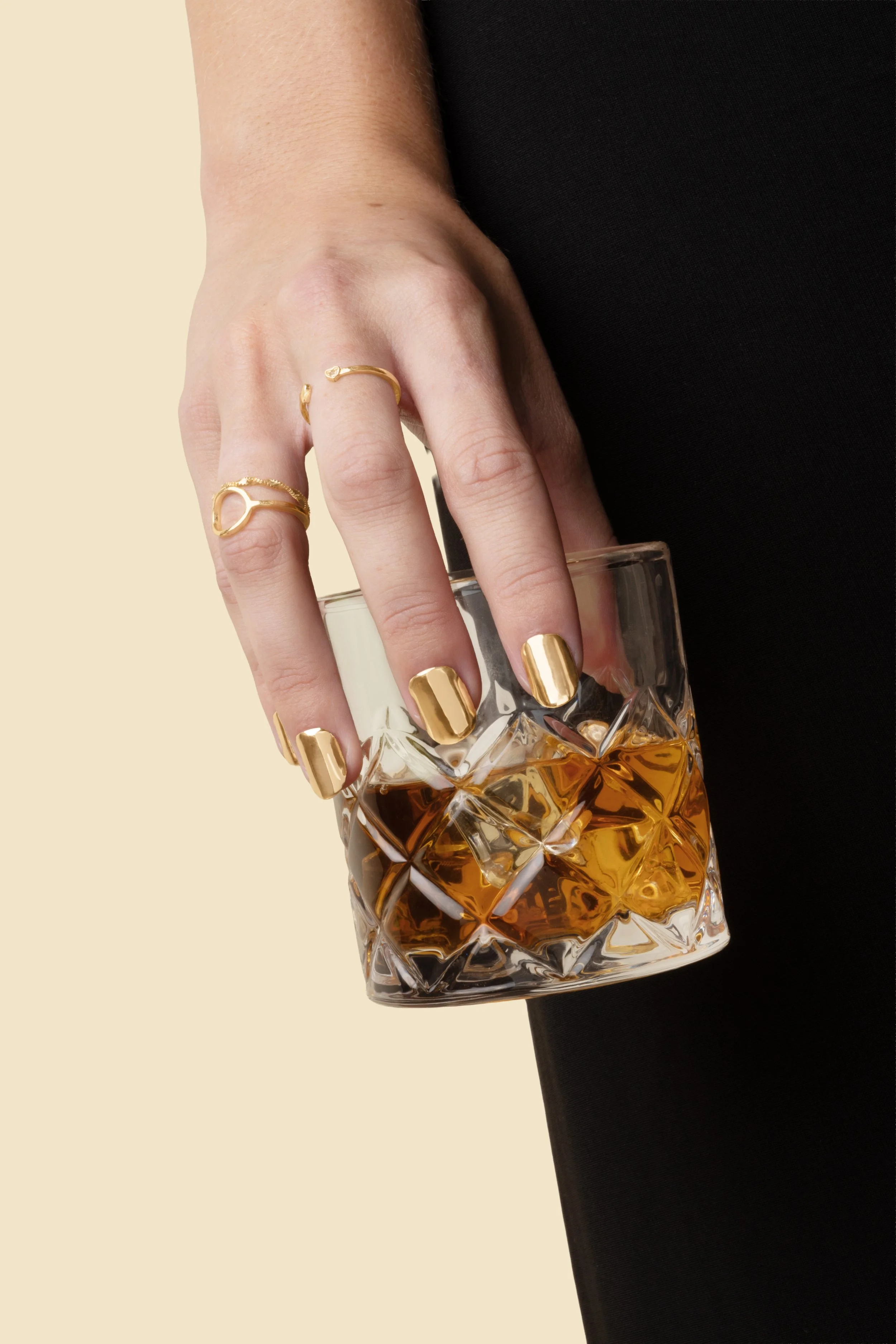 Hand holding a glass wearing 14k gold press-on nails by Stey Gold with gold rings against a beige background