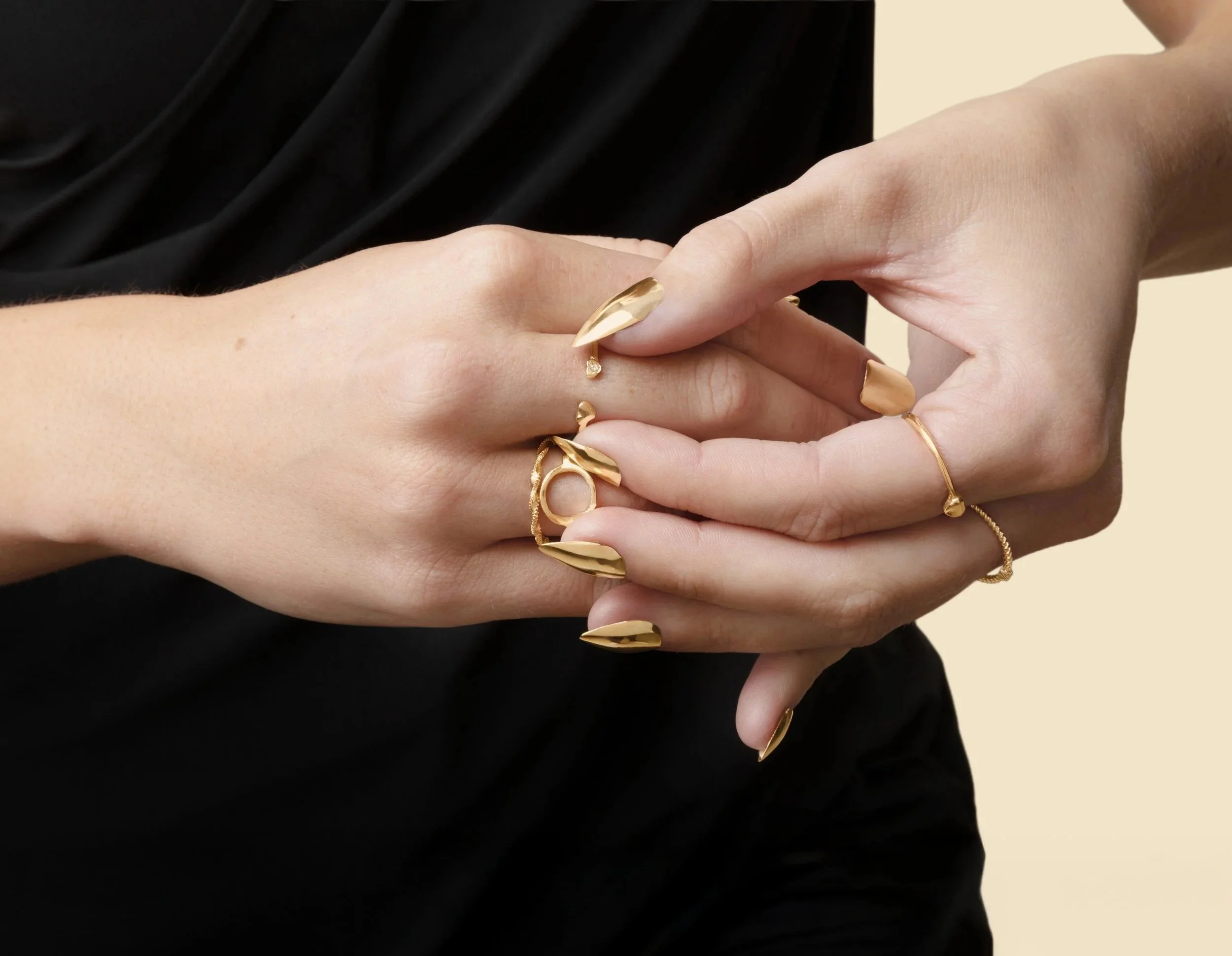 Hands wearing 14k gold press-on nails by Stey Gold with multiple gold rings against a beige background
