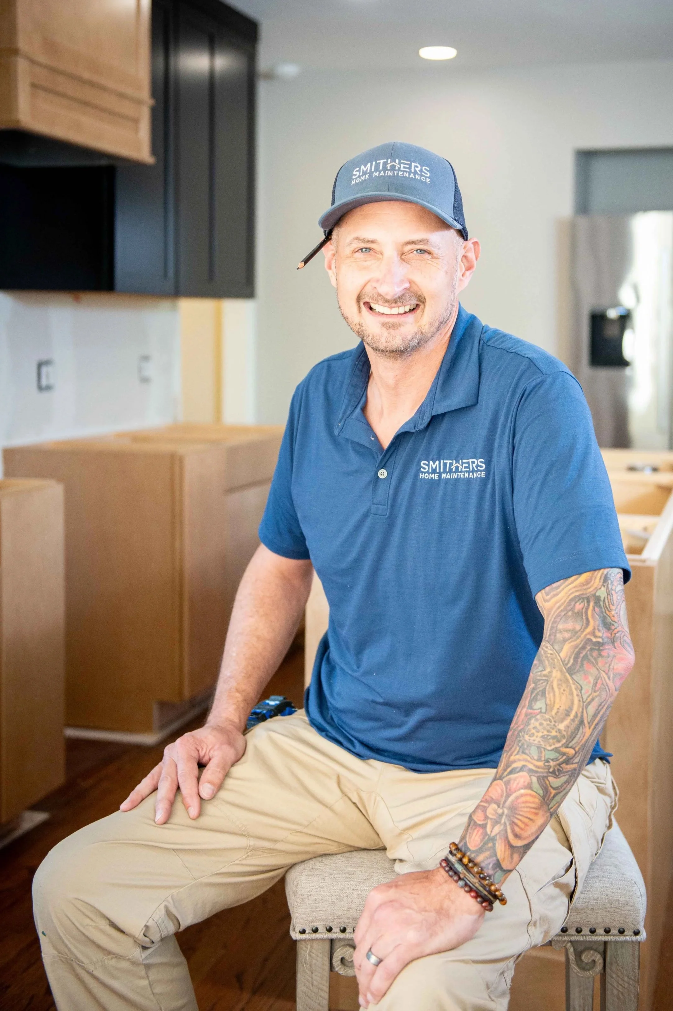 James Smithers in a blue polo shirt and a cap that reads 'Smithers Home Maintenance' is sitting on a beige stool in a home renovation setting, with wooden cabinets and a refrigerator in the background.
