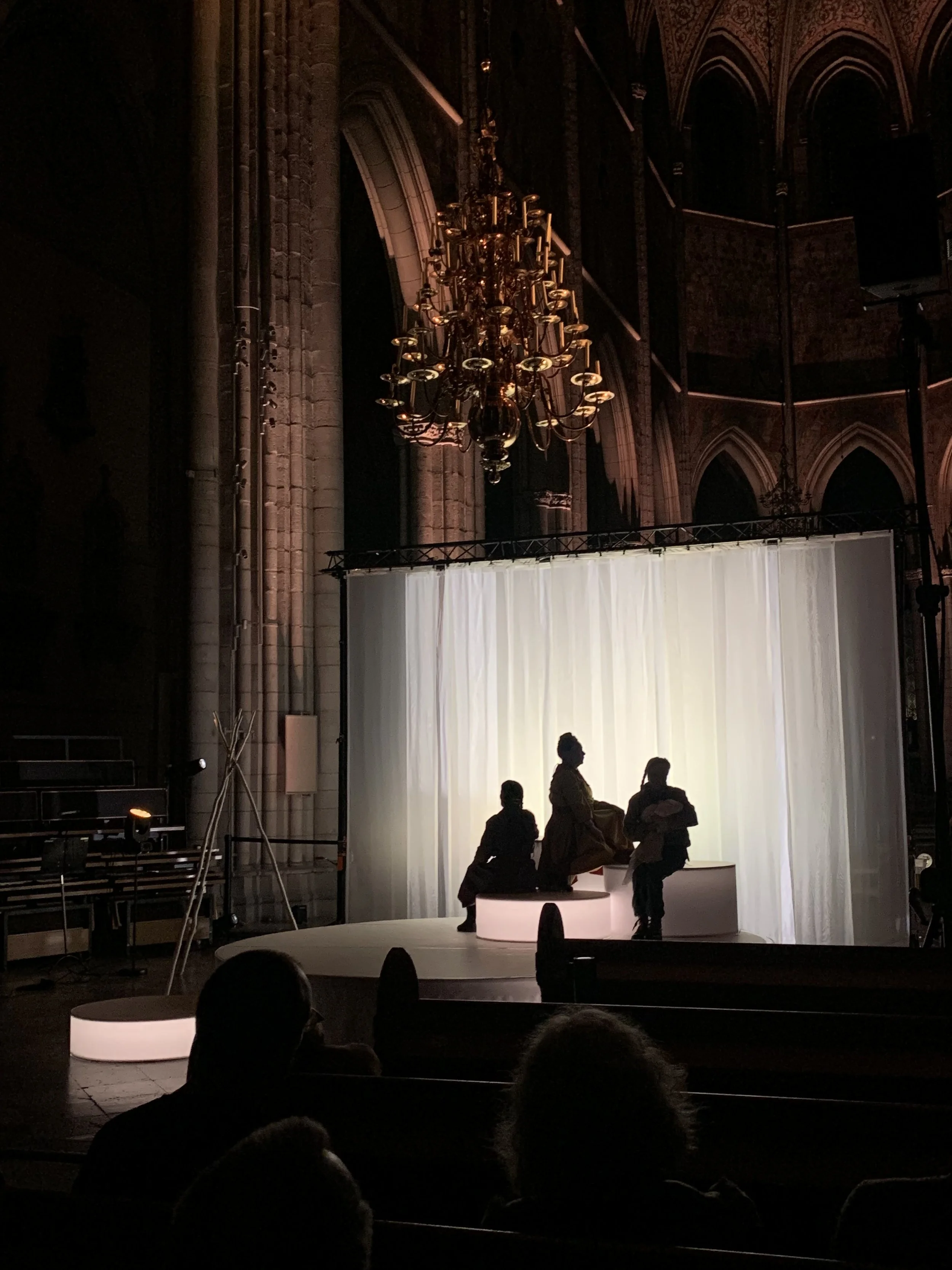 Silhuetter av tre personer på en scen med bakgrund av ljus, inomhus i en stor, gammal byggnad.