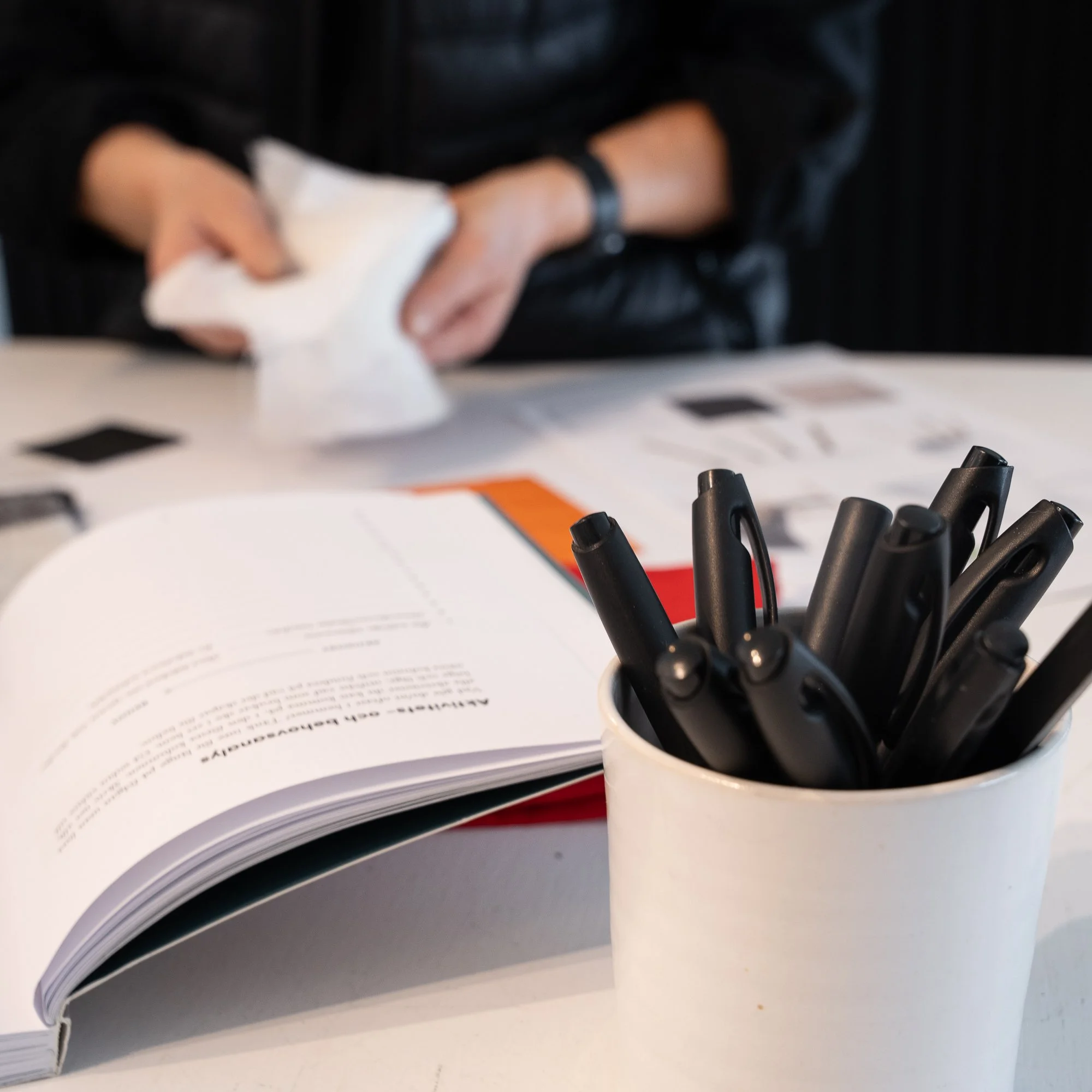 En öppen bok och en penna i en vit kruka på ett bord, med en person i bakgrunden som tittar på tygprover.