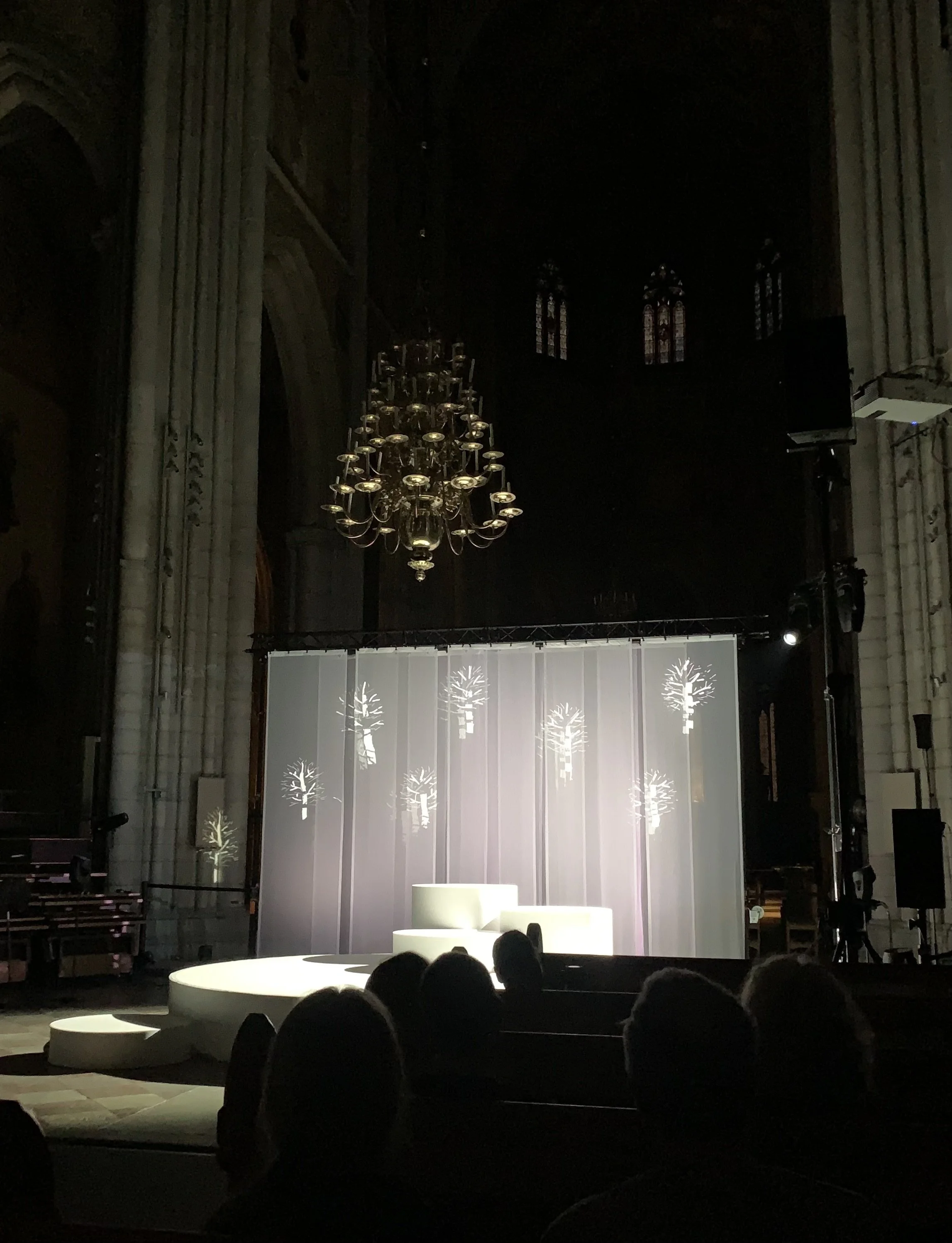En scenuppsättning i en kyrka med ett cirkelformat podium och bakgrund med badliljor. Mörkt utrymme, med en kristallkrona hängande i taket.
