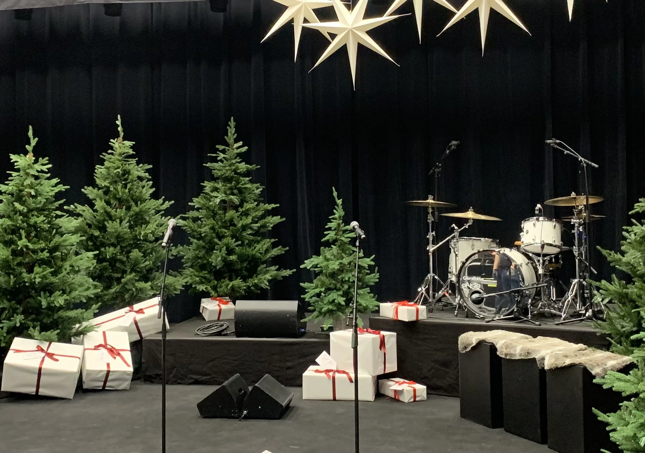 En scen med julgranar, presentaskar med röda band, mikrofoner, ett trumset och en stjärnor hängande ovanför scenen.