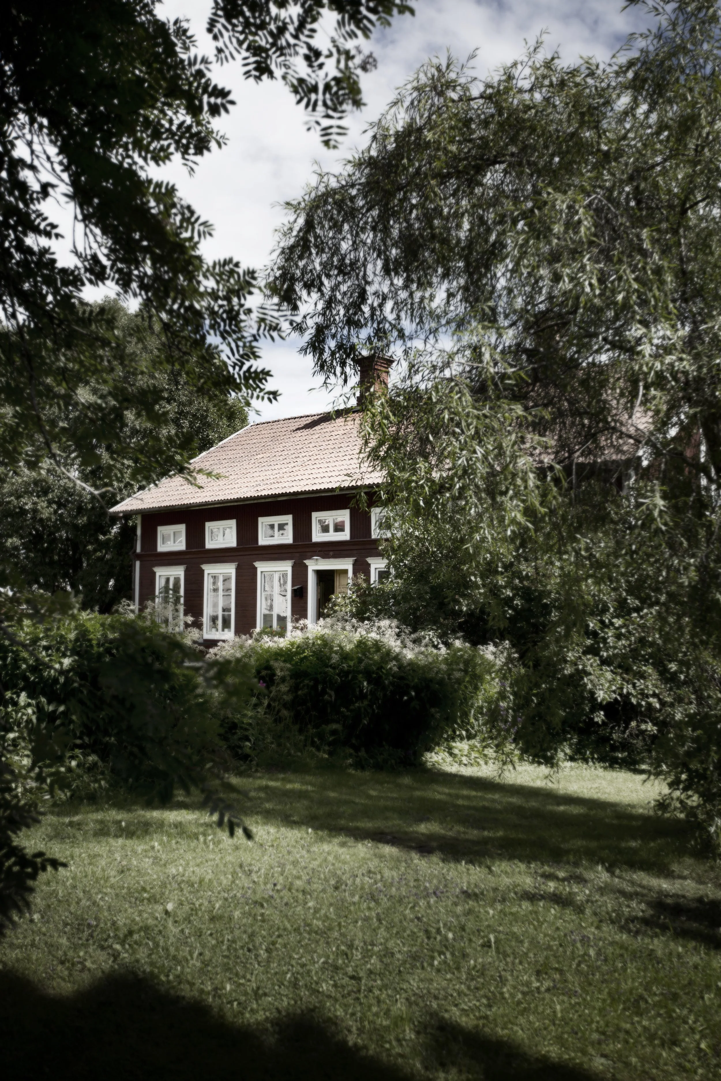 Byhuset, rött hus med vita fönster, omgiven av träd och gräs, sett genom ett träd. Himmel ovan.