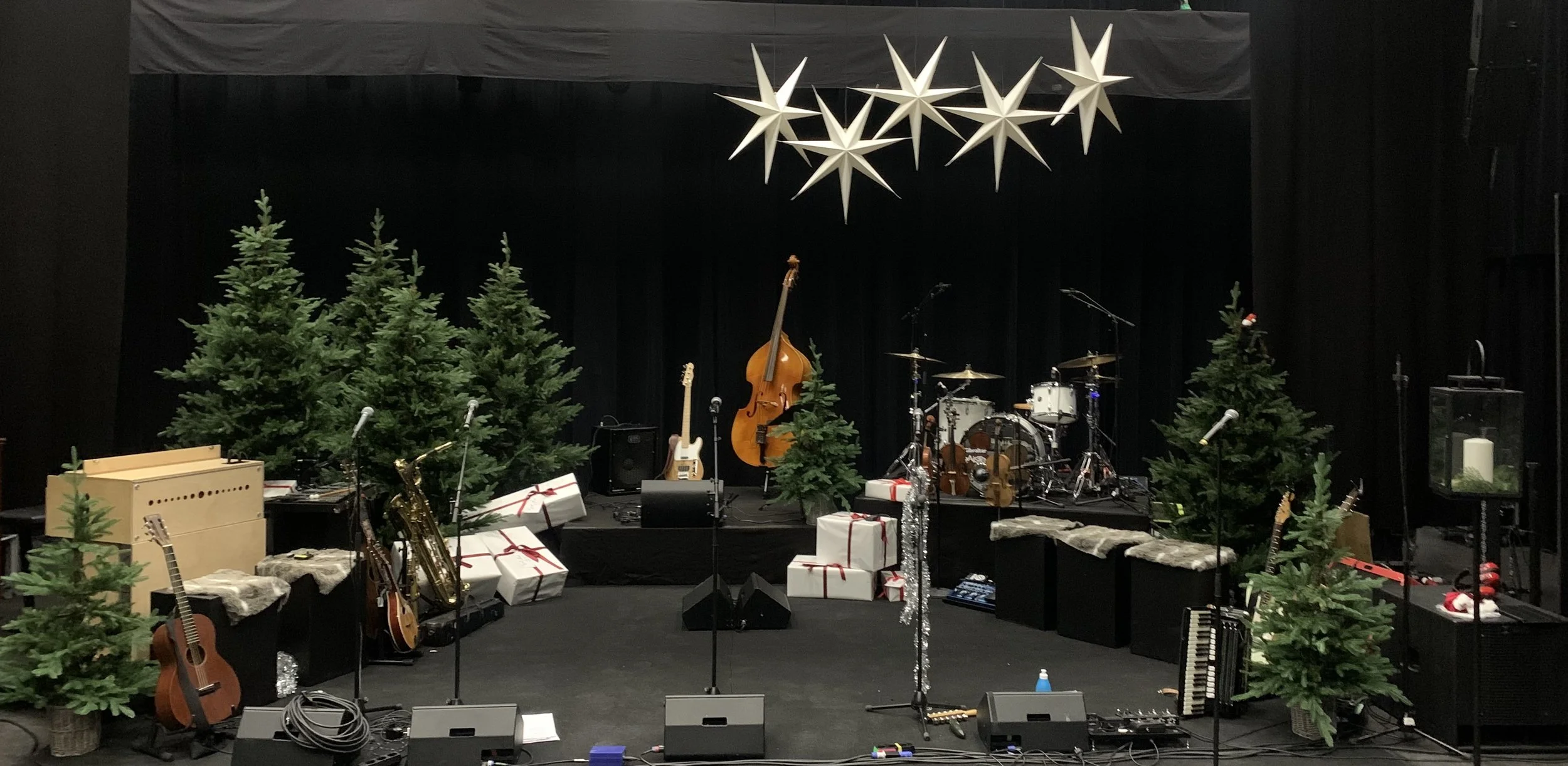 En scen med julgranar, musikinstrument och paket, förberedd för en julkonsert. Hängen av stjärnor över scenen.