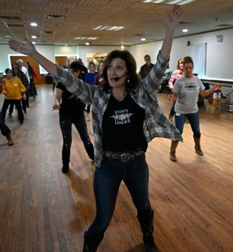 Woman leading a line dance with arms raised, wearing a headset microphone, in a spacious room with multiple people in the background.