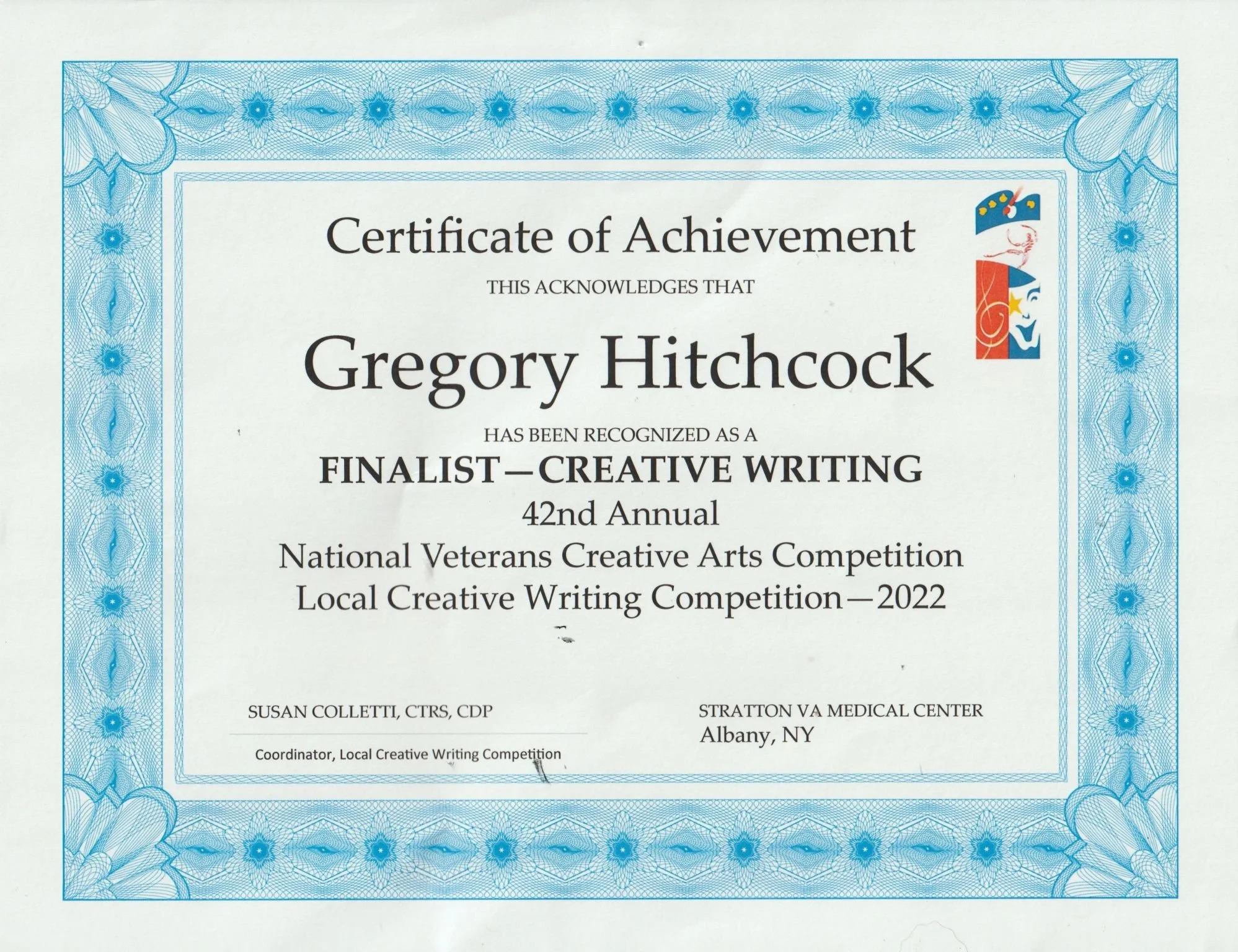 Certificate of Achievement awarded to Gregory Hitchcock for being a finalist in the 42nd Annual National Veterans Creative Arts Competition and Local Creative Writing Competition in 2022, with a decorative blue border and a small colorful emblem in the top right corner.