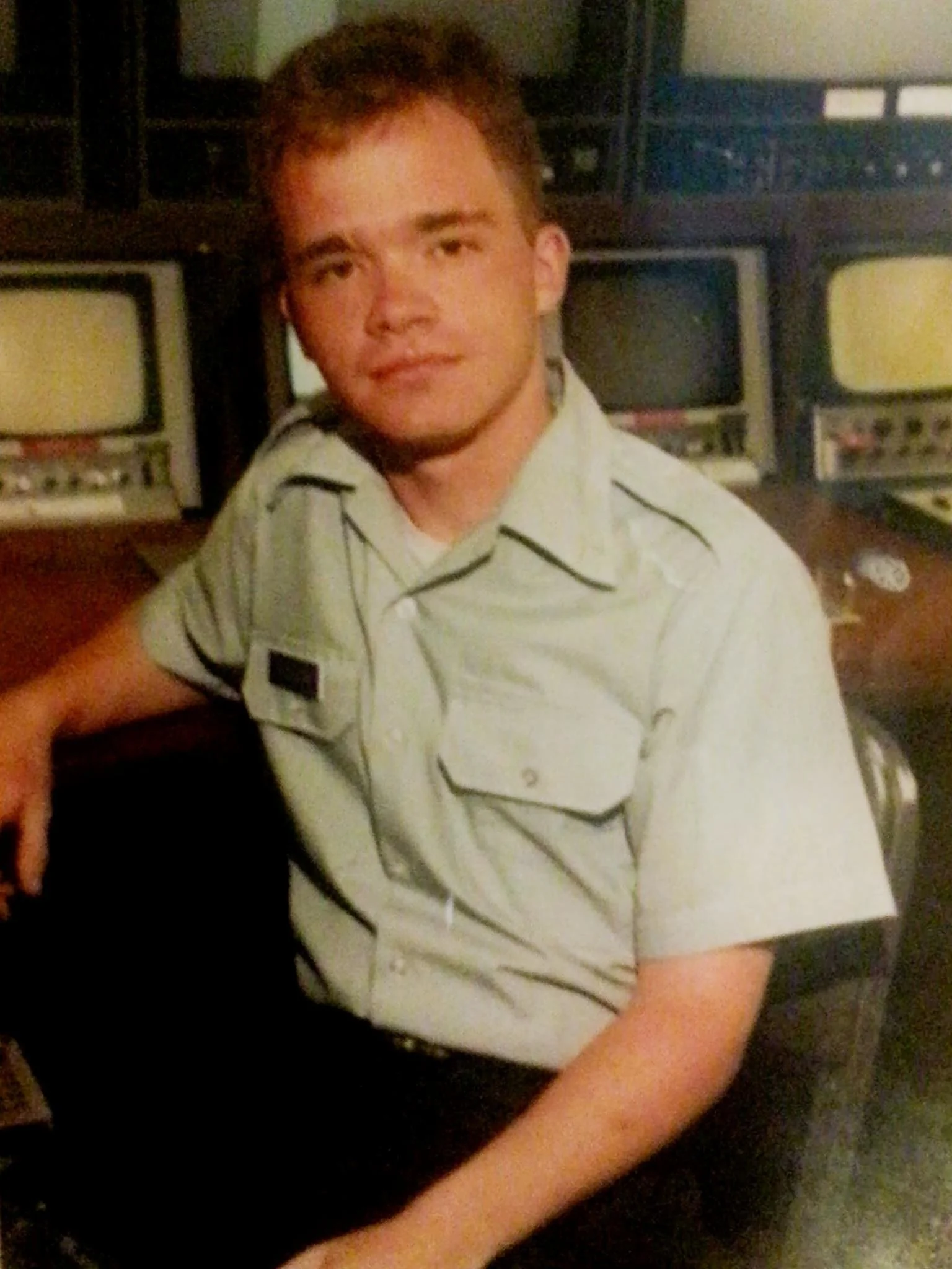 A young man in a light-colored military or police uniform sitting in front of vintage computer monitors.