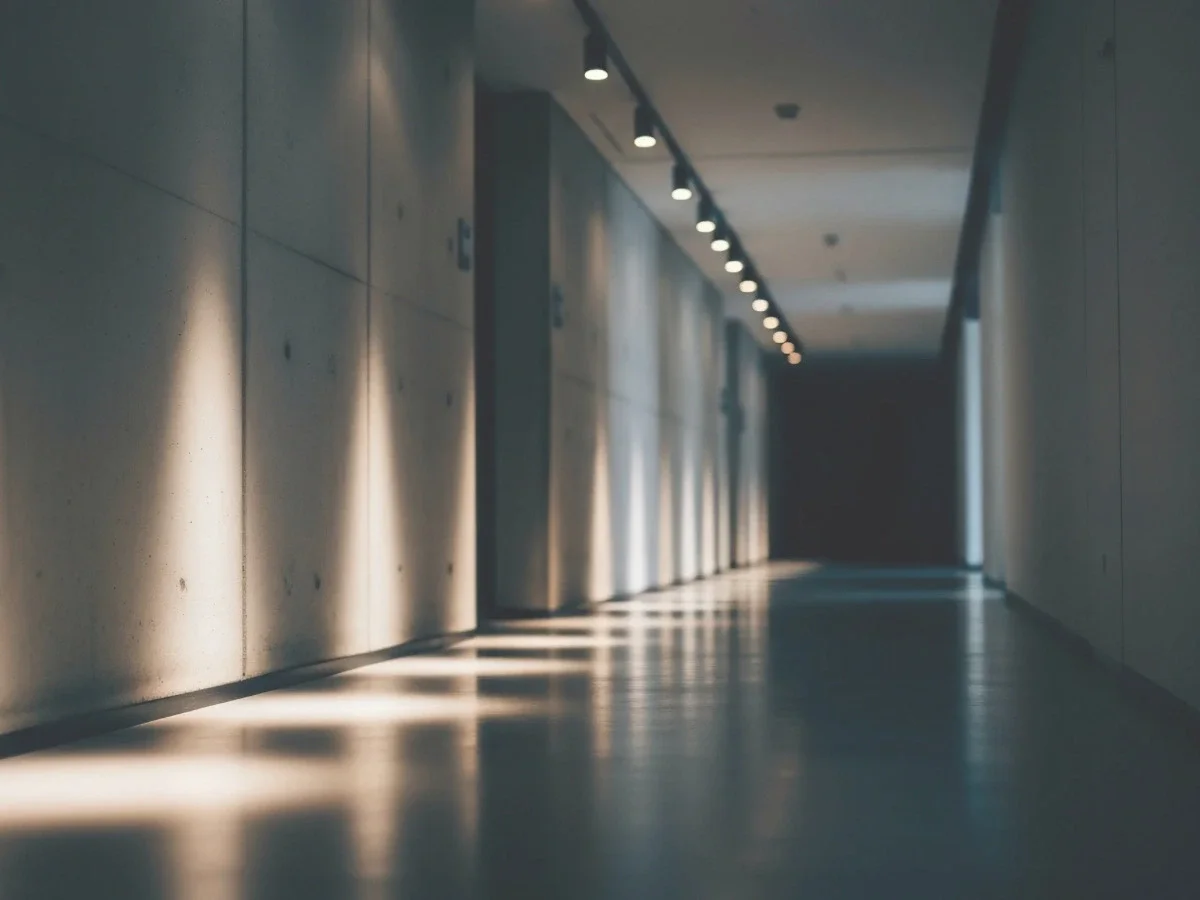 A dimly lit corridor with a repeating sequence of illuminated doorways, photographed in shadow — accompanying the "Practical According to Whom?" section of She The King's essay on desire and self-editing.