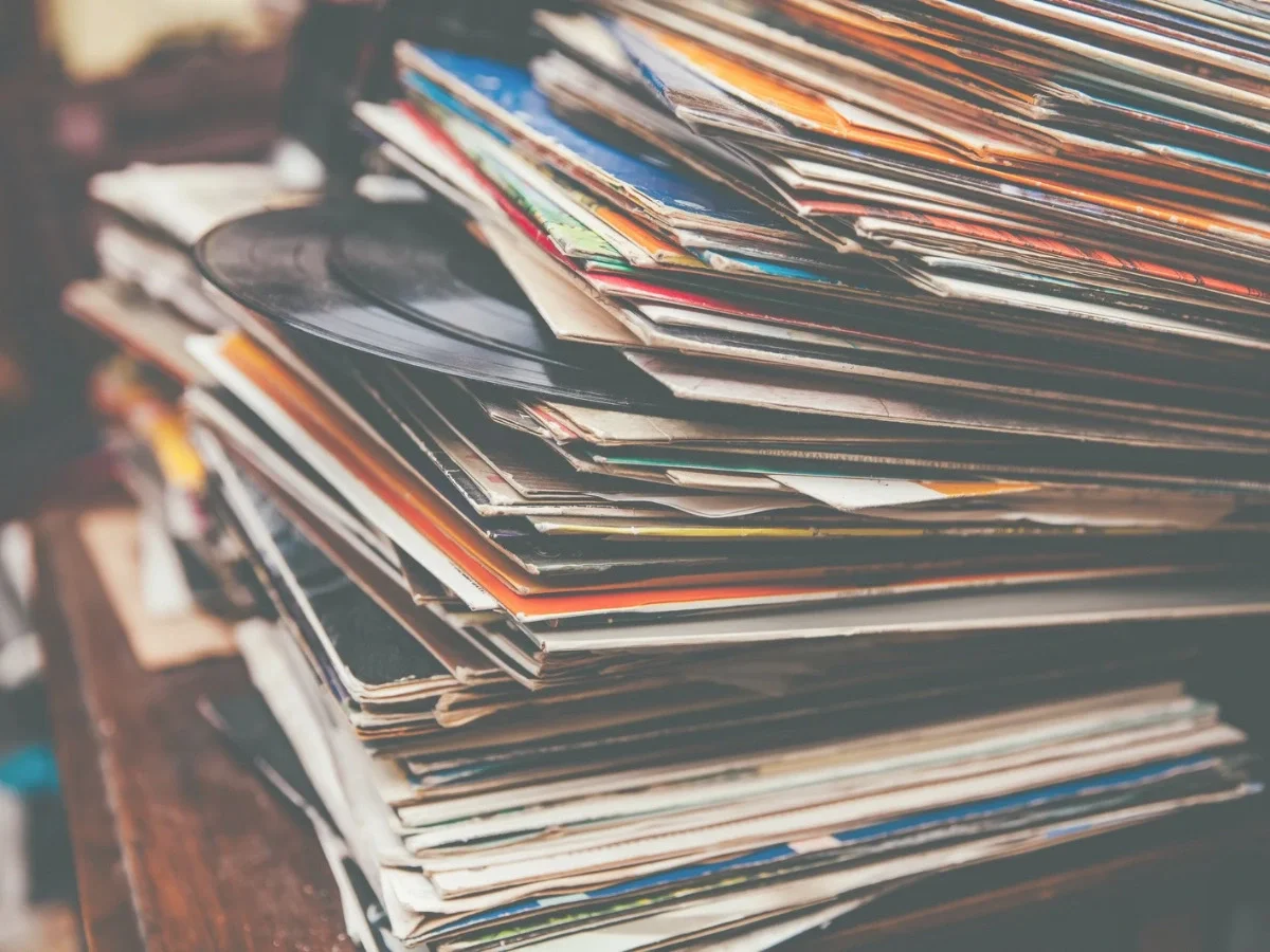 A tall, worn stack of vinyl records and album sleeves in faded color, one LP visibly peeking out—hero image for She The King's essay "When Music Was Something You Could Hold," on the history of recorded music and physical attention.