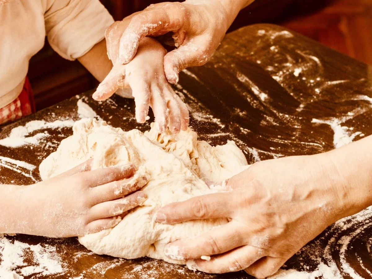 Adult and child's hands kneading bread dough together on a floured wooden table