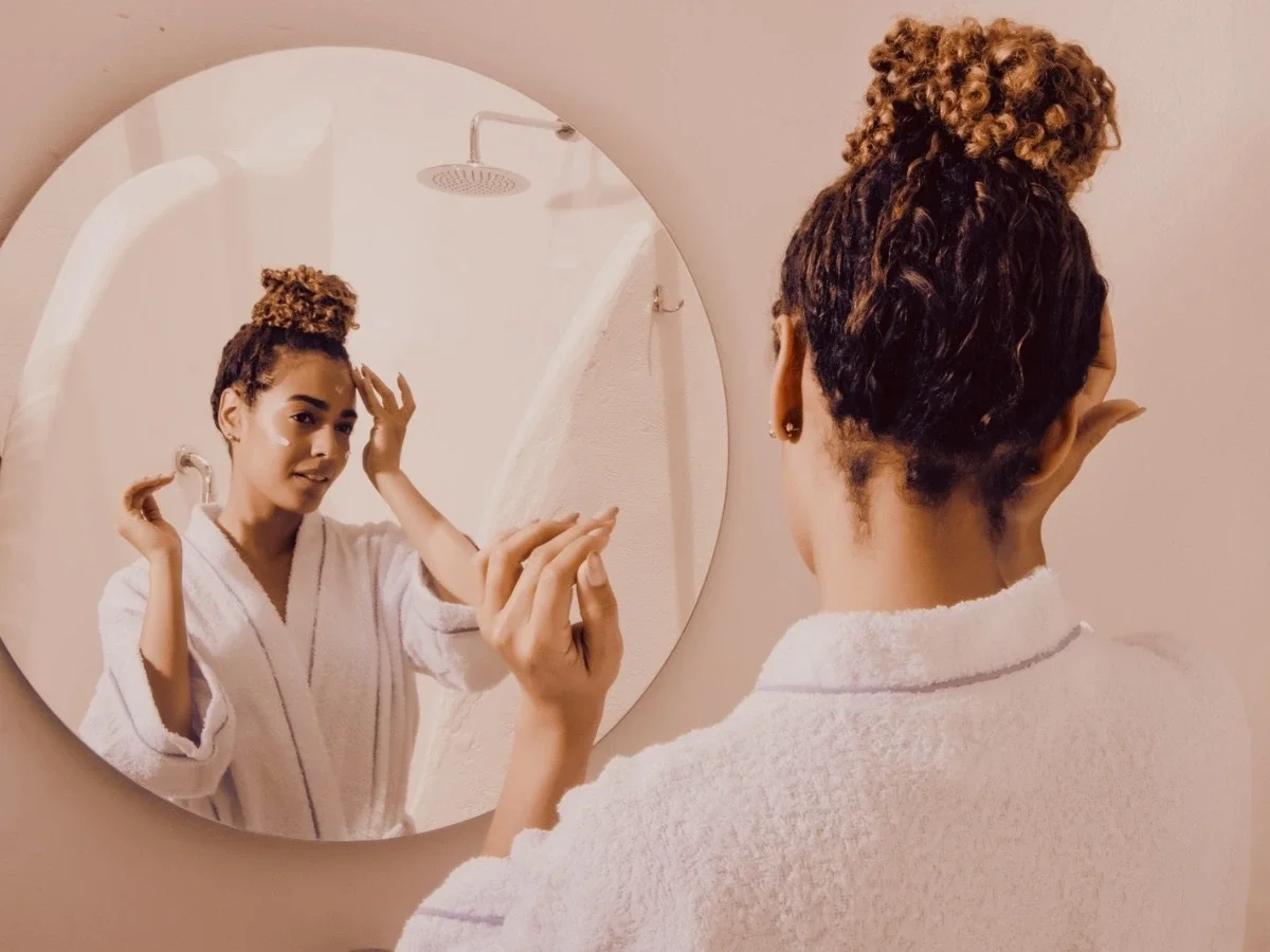Back view of a person in a white robe facing a round bathroom mirror, hands raised mid-routine, with their reflection visible. Blush-toned walls, intimate and unhurried mood.