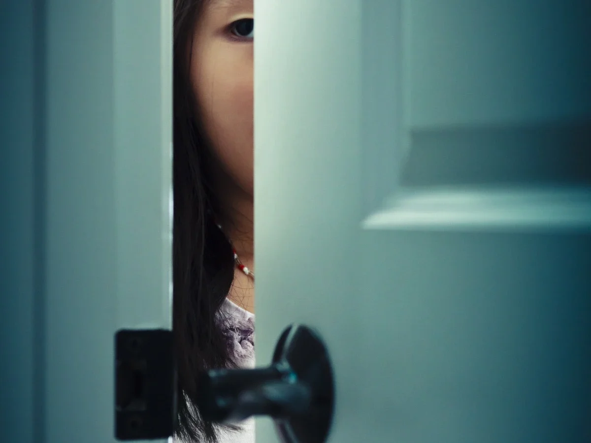 A dark-haired woman's eye visible through a narrow gap in a white door, evoking Ada's refusal to stop looking and her determined search for truth in Minbak by Ela Lee