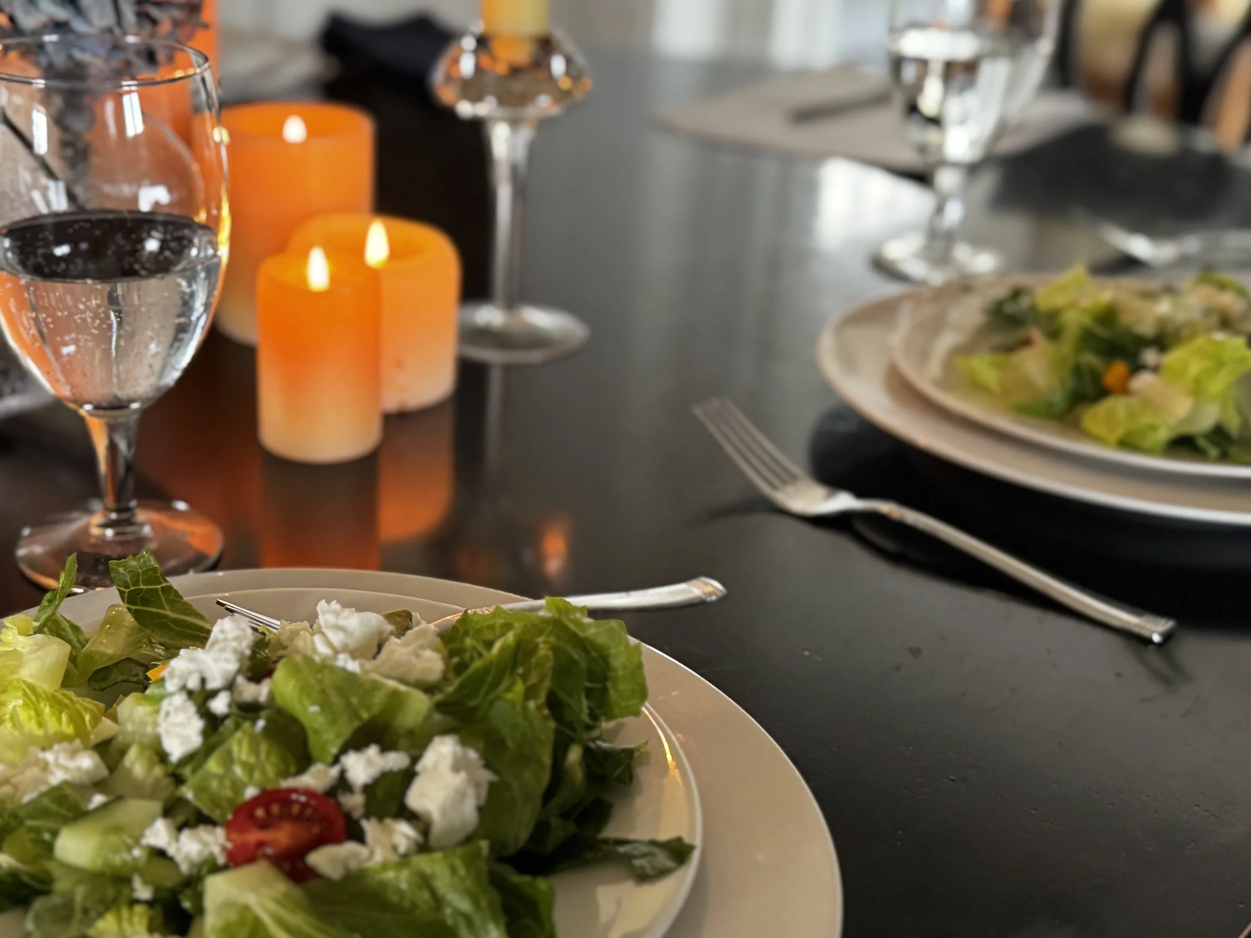 Two place settings with fresh salads and lit pillar candles on a dark dining table — a shared meal set for more than one, representing the emotional practice of eating together.