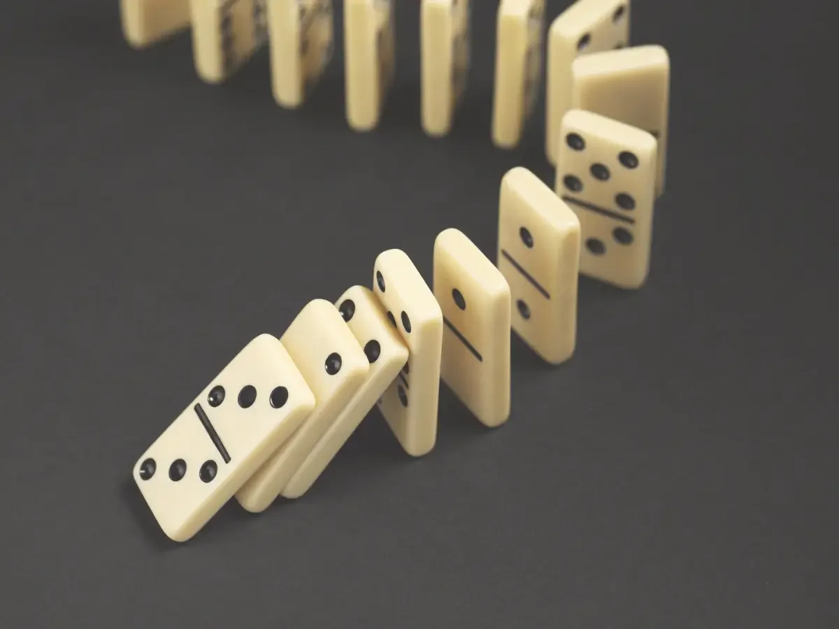 A curved line of ivory dominoes falling in sequence against a dark background — accompanying "The Accumulation of Small Choices," on Michael Corleone's gradual transformation in She The King's essay "When the Villain Feels Familiar.