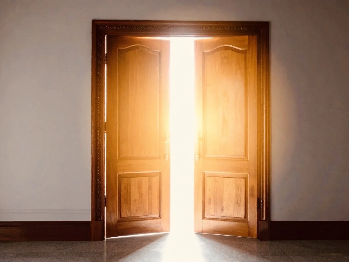 Two tall ornate wooden doors stand open in an empty room, flooding the quiet interior with intense warm golden light from beyond the threshold. The space ahead remains unseen.