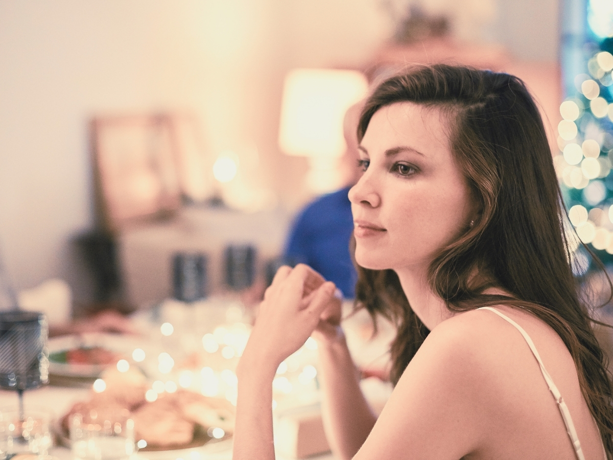 Woman at a lit dinner table, present among others but looking away, lost in thought