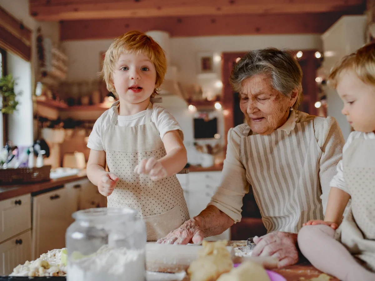 A grandmother kneads dough alongside her young grandchildren in a warm, rustic kitchen, illustrating the everyday traditions passed down through generations before a diaspora traveler ever visits the homeland.