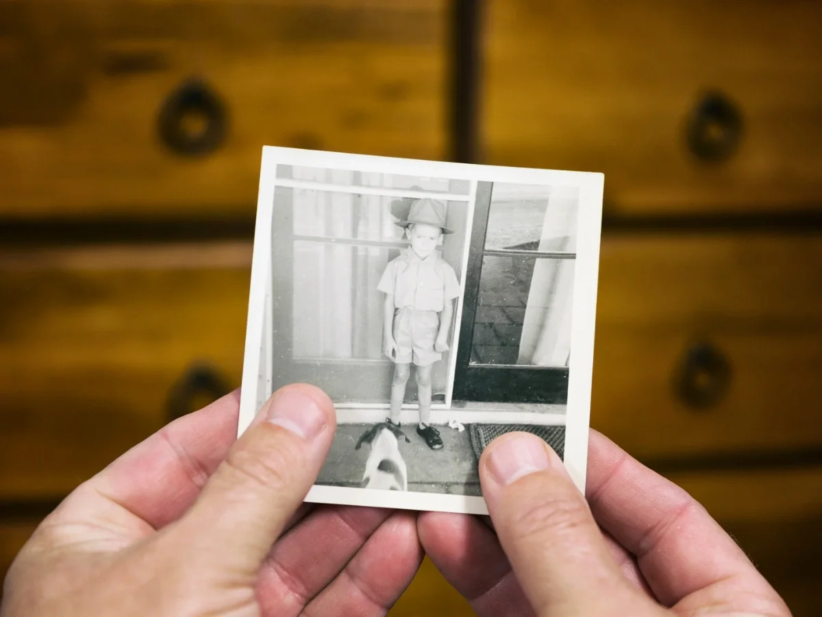 Hands holding a small vintage black-and-white photograph of a young boy standing in a doorway, evoking the inherited family memories diaspora travelers carry into unfamiliar places.