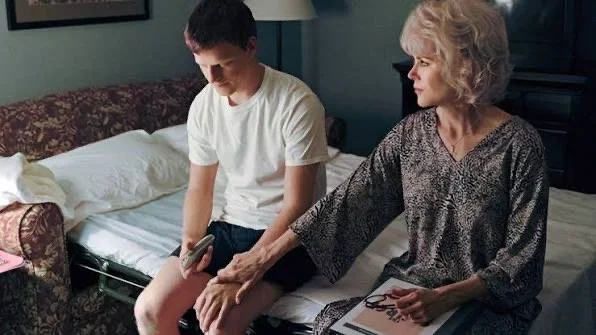 A young man sits tensely on a bed beside an older woman who gently touches his hand, reflecting themes of family tension, rejection, and emotional conflict in queer storytelling.