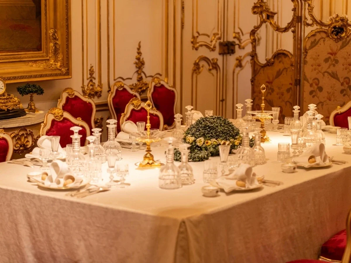An elaborate historical dining room with gold panelling, red velvet chairs, and a formally set table crowded with crystal decanters, white porcelain, and gold centrepieces. No guests present. The table as an artifact of wealth and social hierarchy.