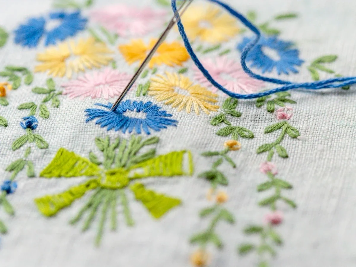 A macro close-up of floral embroidery in progress on white linen — a needle mid-pull trailing blue thread through dense, multicolored stitches in cobalt, yellow, pink, and green. The sophistication of the work fills the frame.