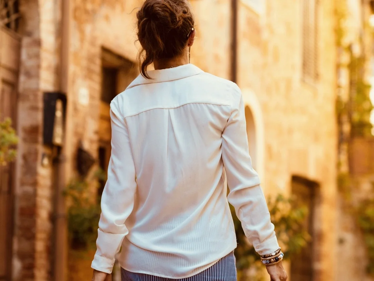 A woman in a white blouse and stacked gold bracelets walks away from camera down a narrow stone street bathed in warm amber light. Shot from behind, her destination remains unrevealed.
