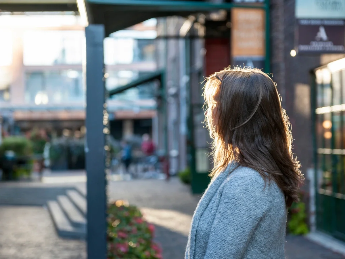 A woman paused mid-stride on a sunlit street, hair lit from behind, evoking the quiet uncertainty of becoming someone new