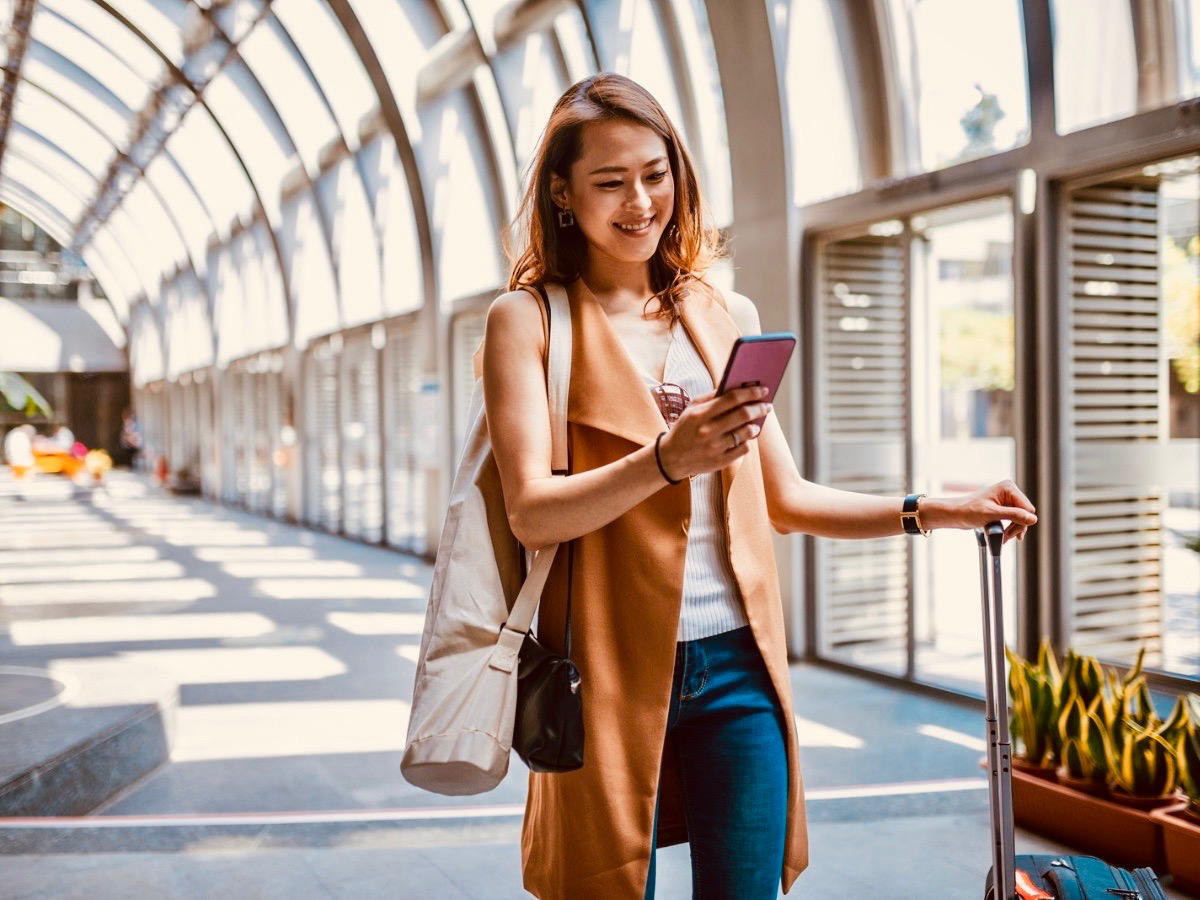 A woman smiling at her phone while pulling luggage through a bright airport terminal