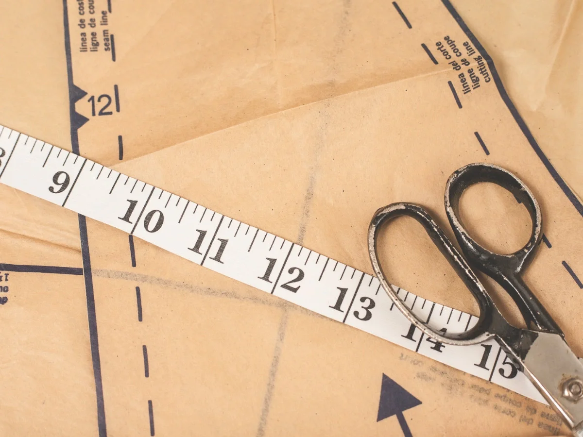 A vintage tape measure and scissors laid across a paper sewing pattern marked with seam lines and numbers — accompanying "Learning to Make It Palatable," on self-editing and alteration in She The King's Apfel essay.
