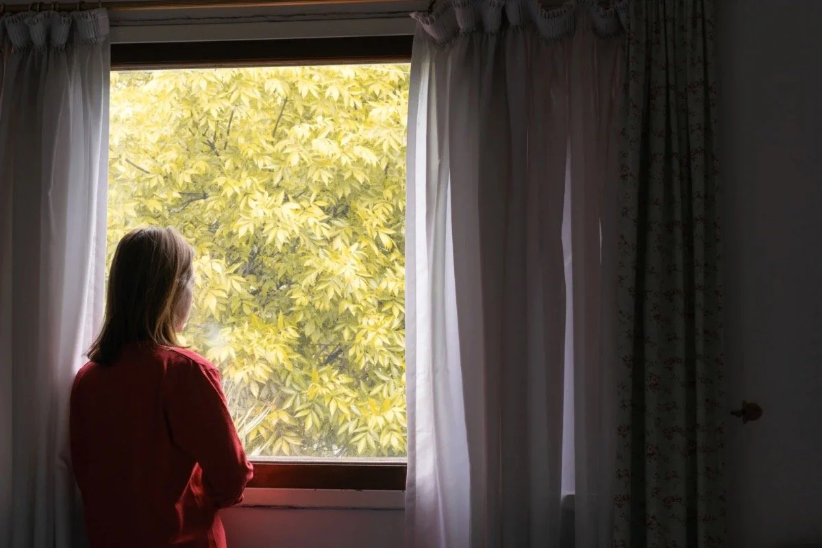 A woman stands quietly by a window, autumn light filtering through sheer curtains.