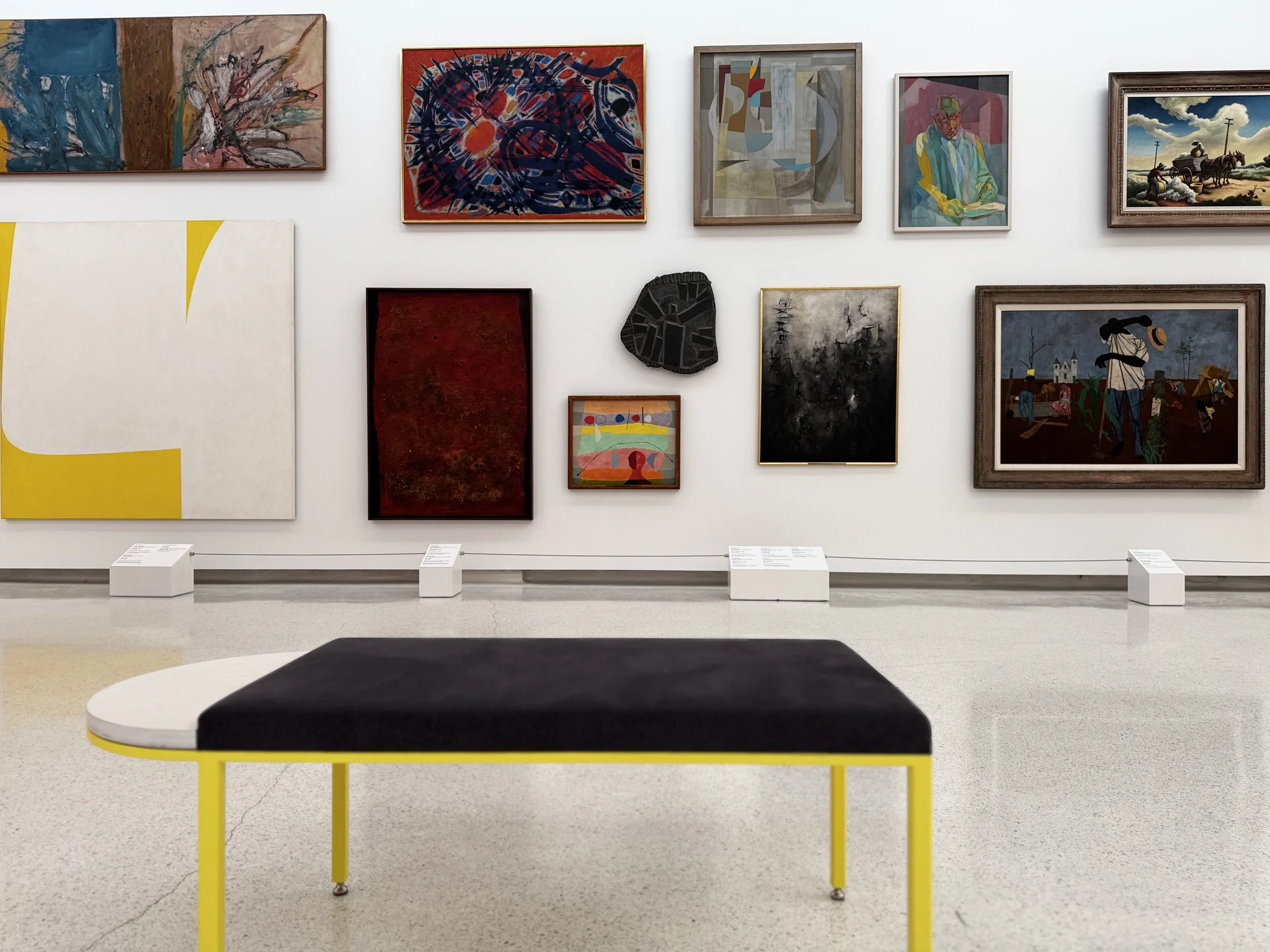 A museum gallery with a black-cushioned bench in the foreground and a wall of paintings behind it — an empty space for quiet presence
