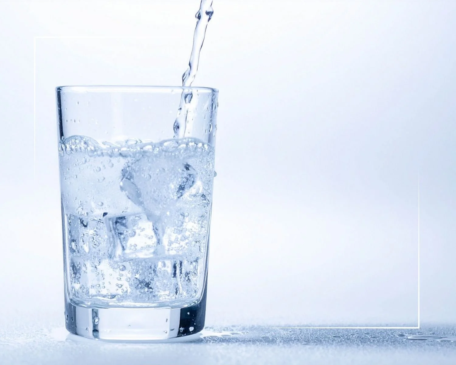 A glass of cold water with ice cubes being filled from above, creating bubbles and droplets, set against a light blue background.