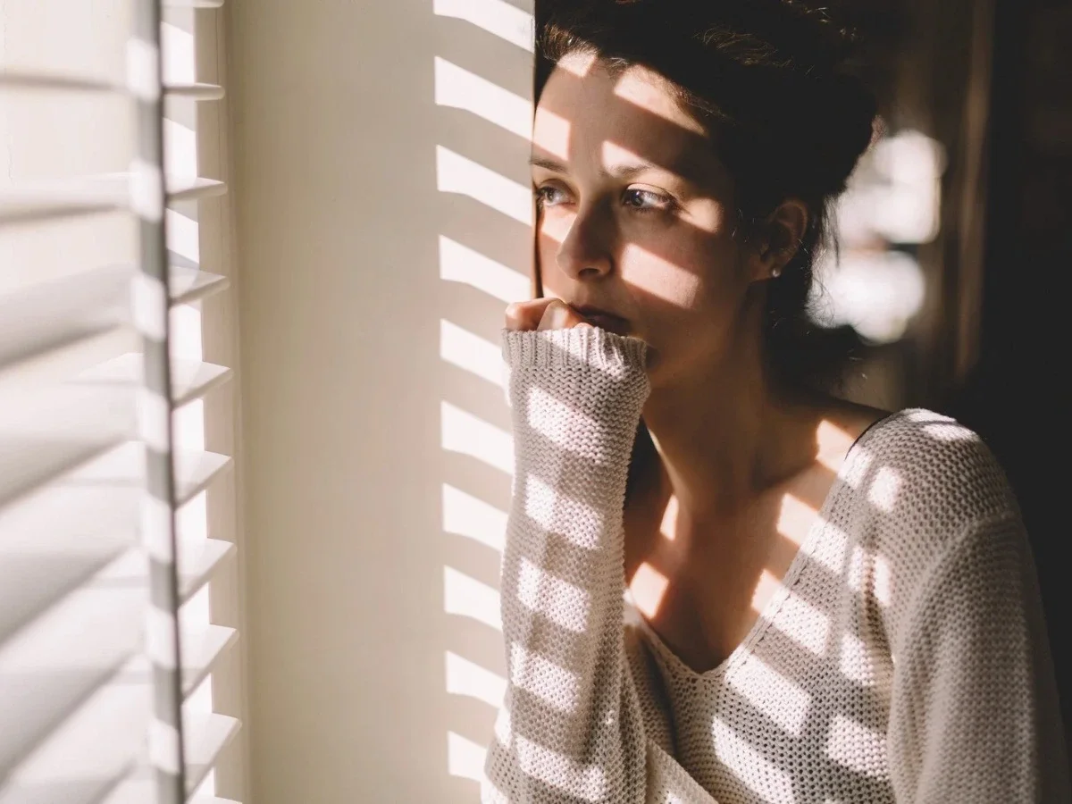 A pensive woman leaning against window blinds, her face and sweater crossed by stripes of soft light — a quiet moment of inner contemplation.
