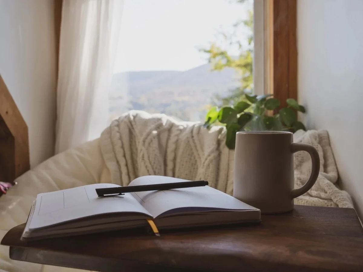 An open journal with blank pages and a pen laid across it sits on a warm wooden surface beside an untouched mug. A mountain view fills the window behind an empty chair draped in a cream knit throw.