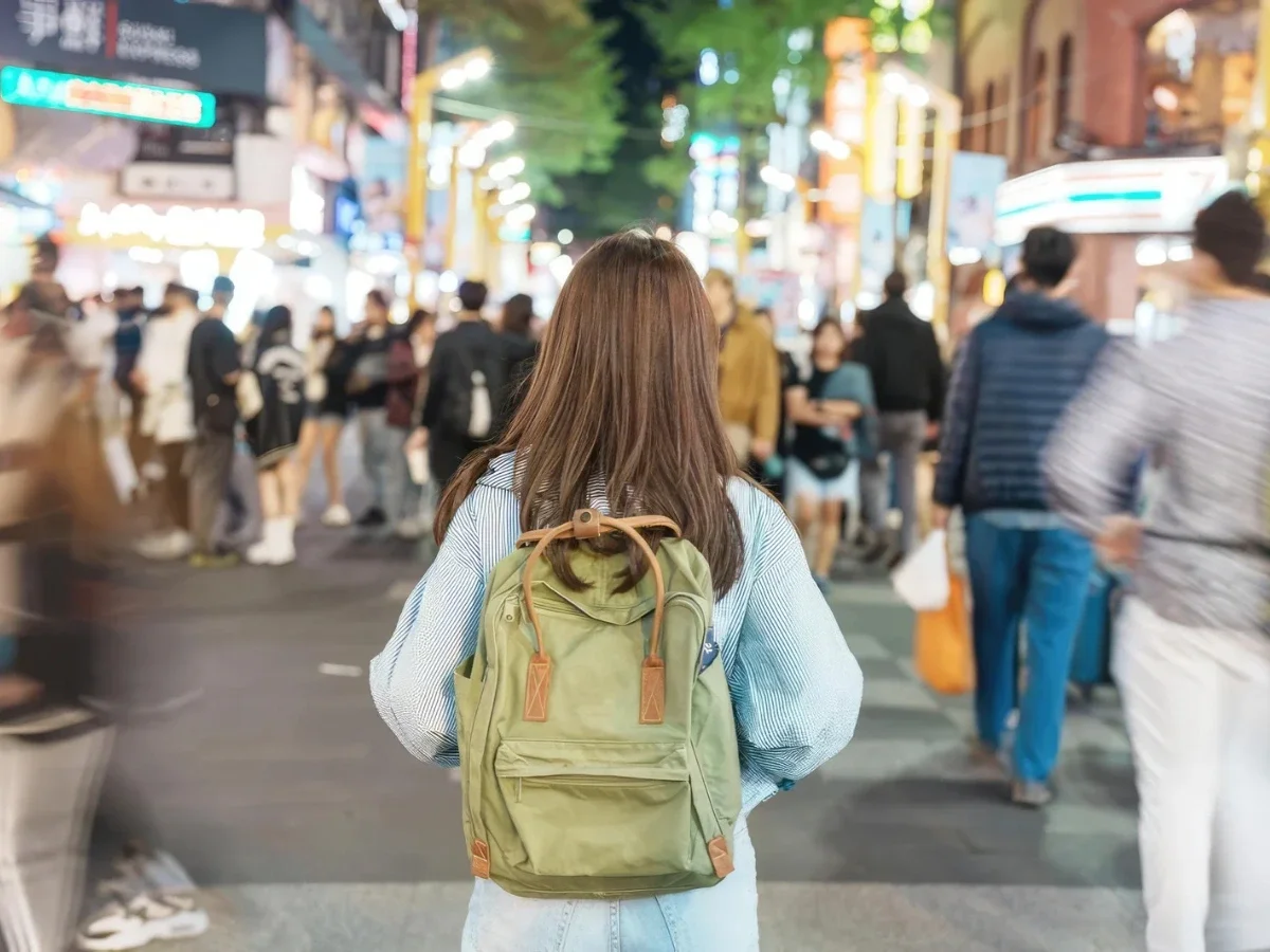 A woman standing still on a crowded night street as blurred crowds move around her, evoking the isolation of a language barrier