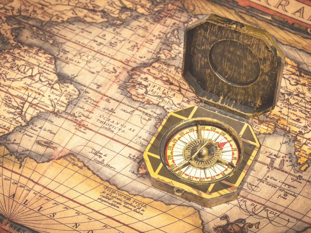 An open brass compass resting on a hand-drawn antique world map showing coastline and ocean routes — illustrating "Information Before Instruction," the section of She The King's essay on desire as a signal rather than a command.