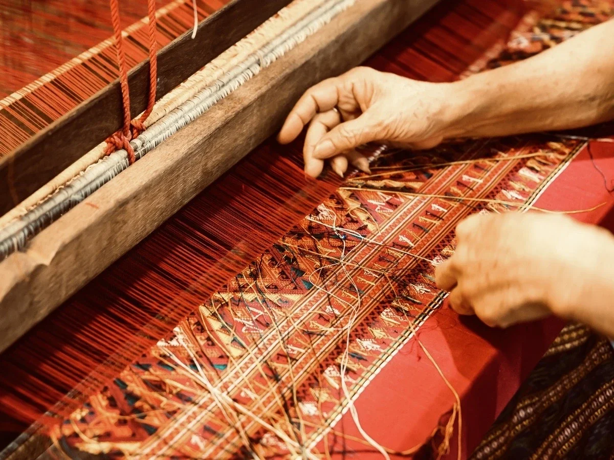 Close-up of weathered hands working a traditional loom, weaving a red and yellow patterned textile by hand