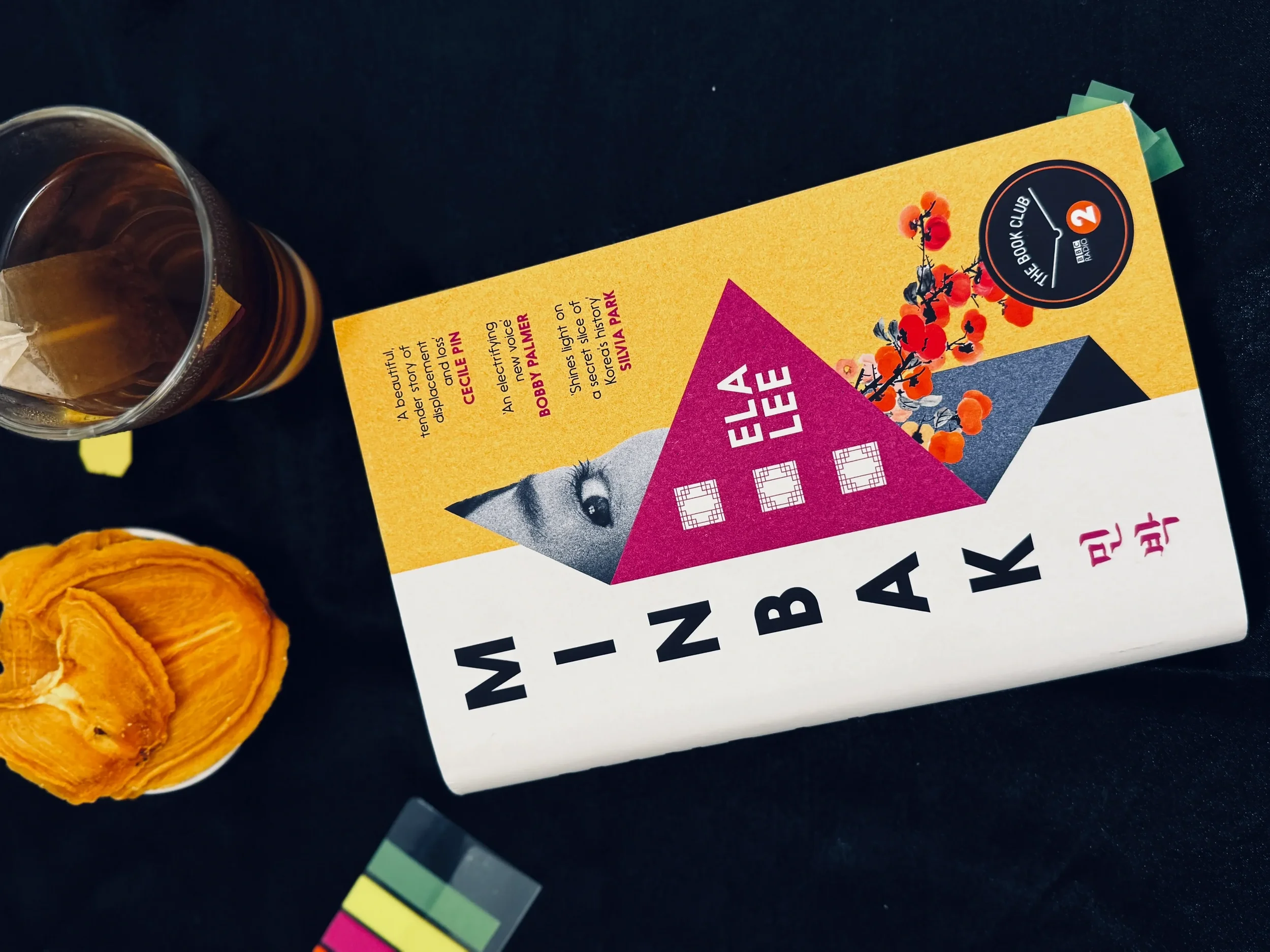 Flat lay of Minbak by Ela Lee lying on a dark surface with sticky note reading tabs, a glass of tea and a persimmon-shaped cookie beside it, evoking the intimate reading experience of this BBC Radio 2 Book Club pick