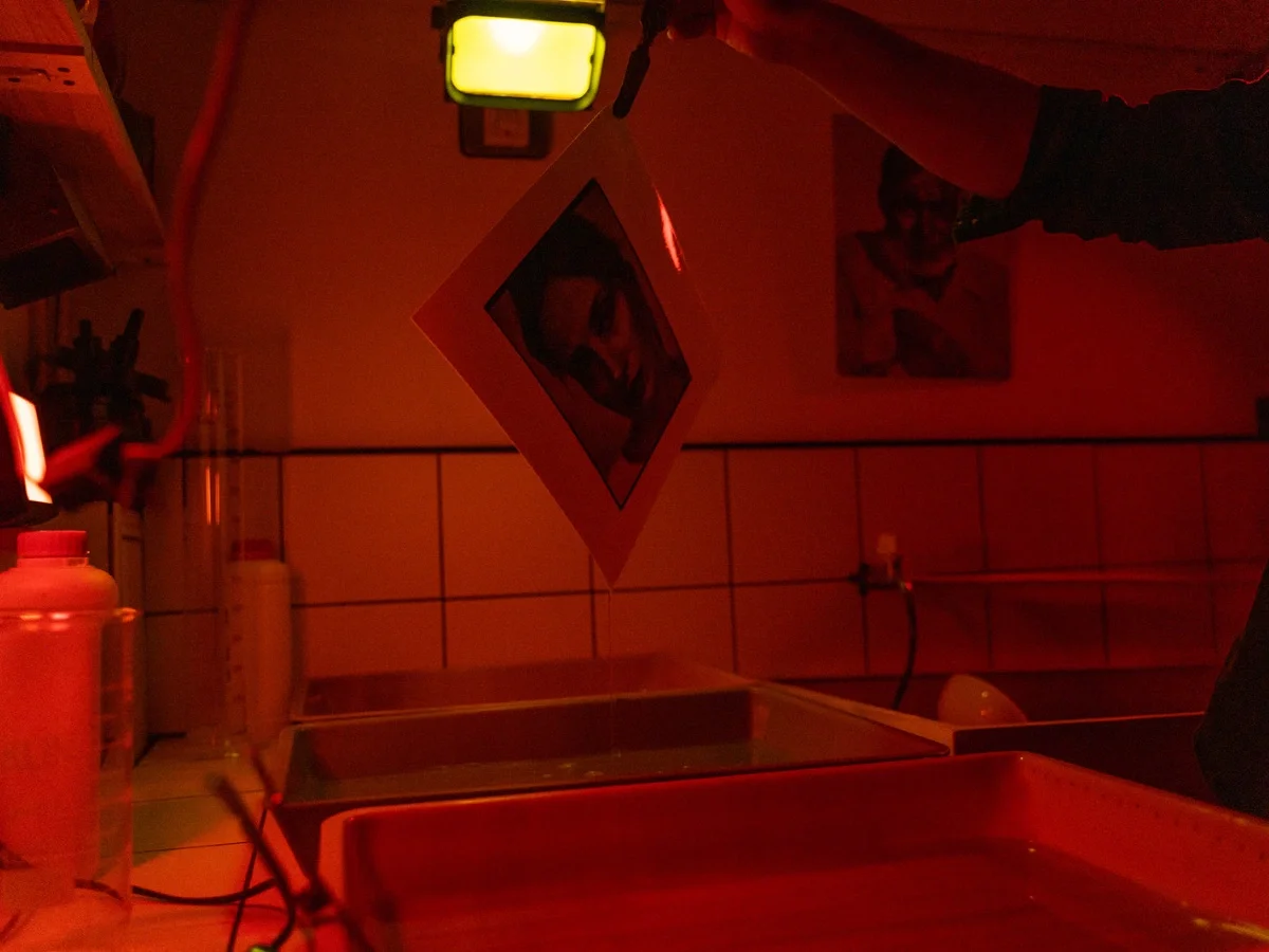 A photographic print hanging to dry above chemical trays in a red-lit darkroom, a hand adjusting the light above