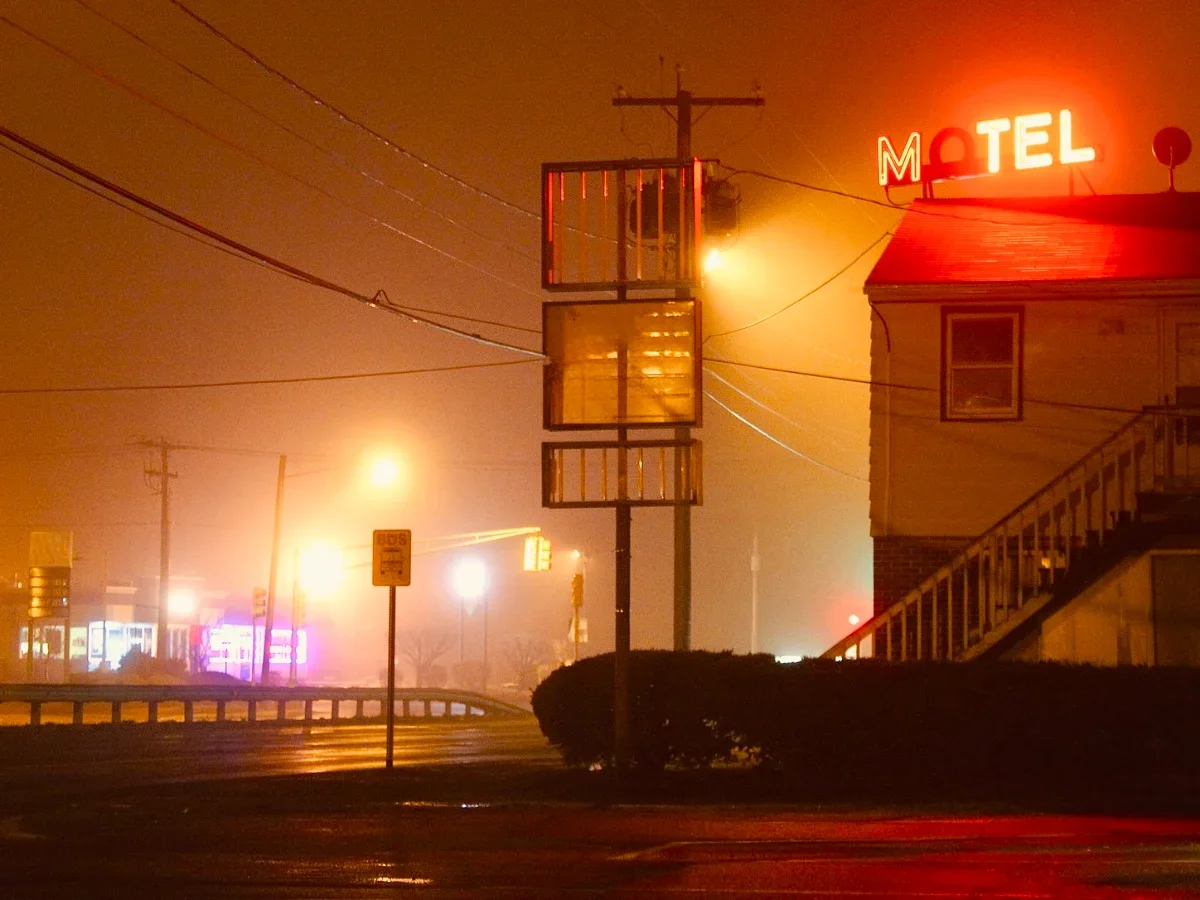 Glowing red neon motel sign against a foggy orange night sky on a wet roadside