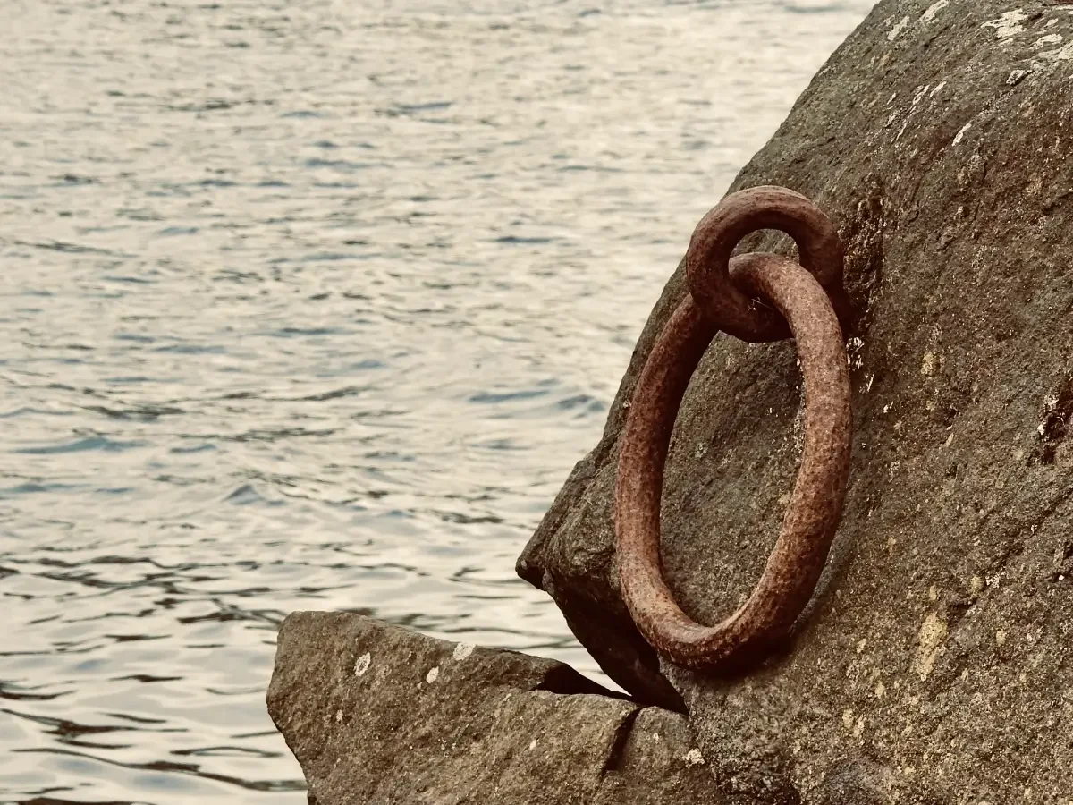 A weathered iron mooring ring fixed into coastal rock beside the sea, suggesting the maritime world of The Pirate Queen.