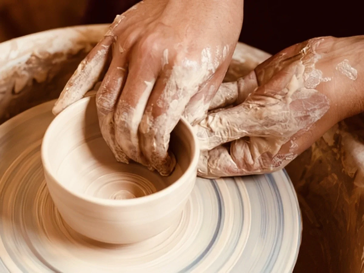 Two clay-covered hands shape a ceramic vessel on a spinning pottery wheel, seen from above in warm studio light. The form is mid-process — the maker's touch still visible in the clay.